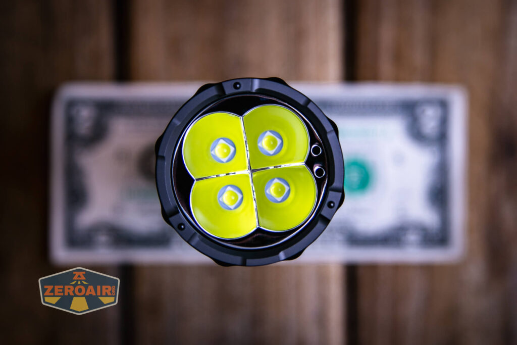 A close-up, top-down view of a four-LED flashlight head placed on top of a one-dollar bill on a wooden surface, with a ZeroAir logo in the bottom left corner.