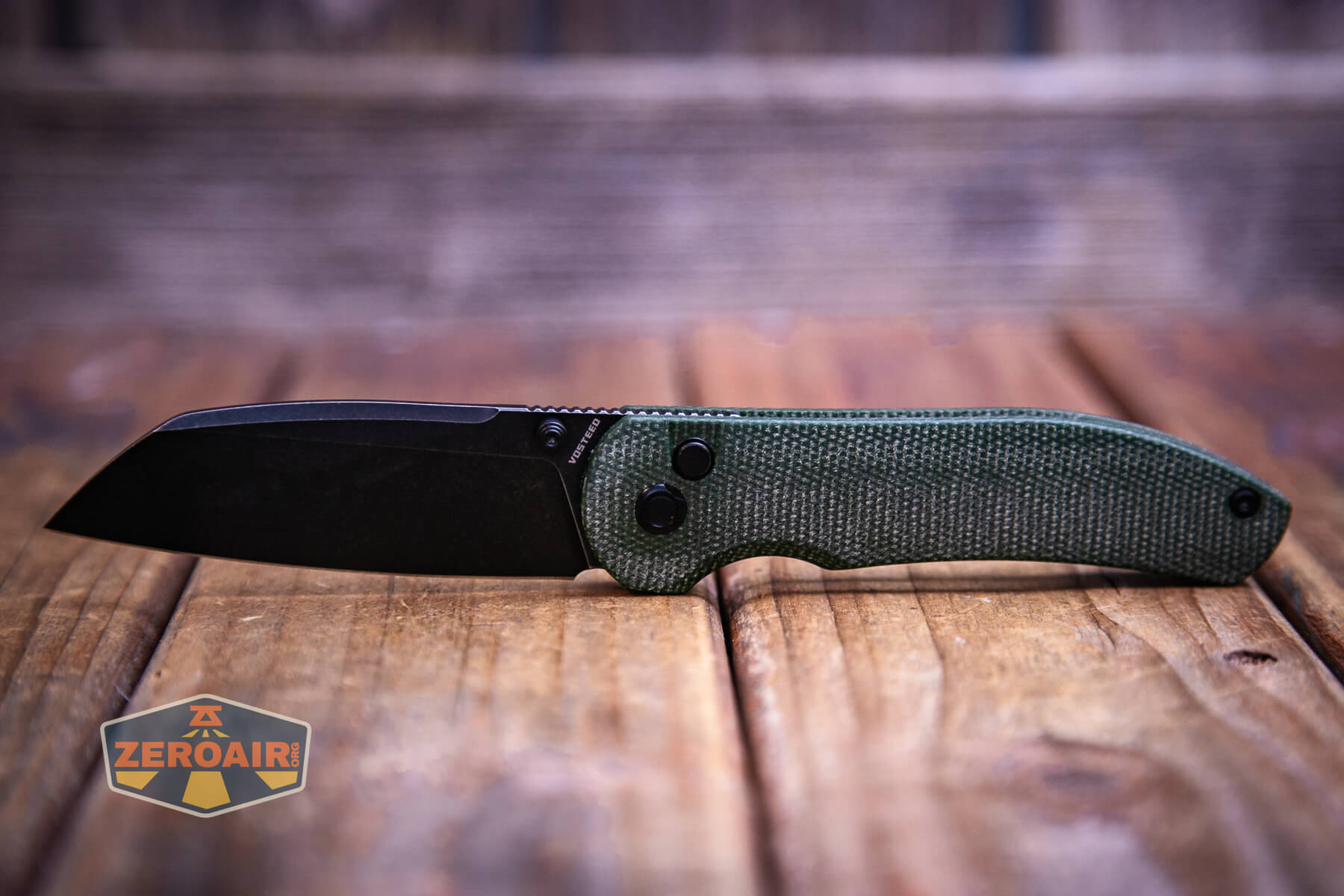 A Vosteed Thornton Trek lock knife with a black blade and a textured green handle is displayed on a wooden surface. The background is blurred wood, and a Zeroair logo appears in the lower left corner.