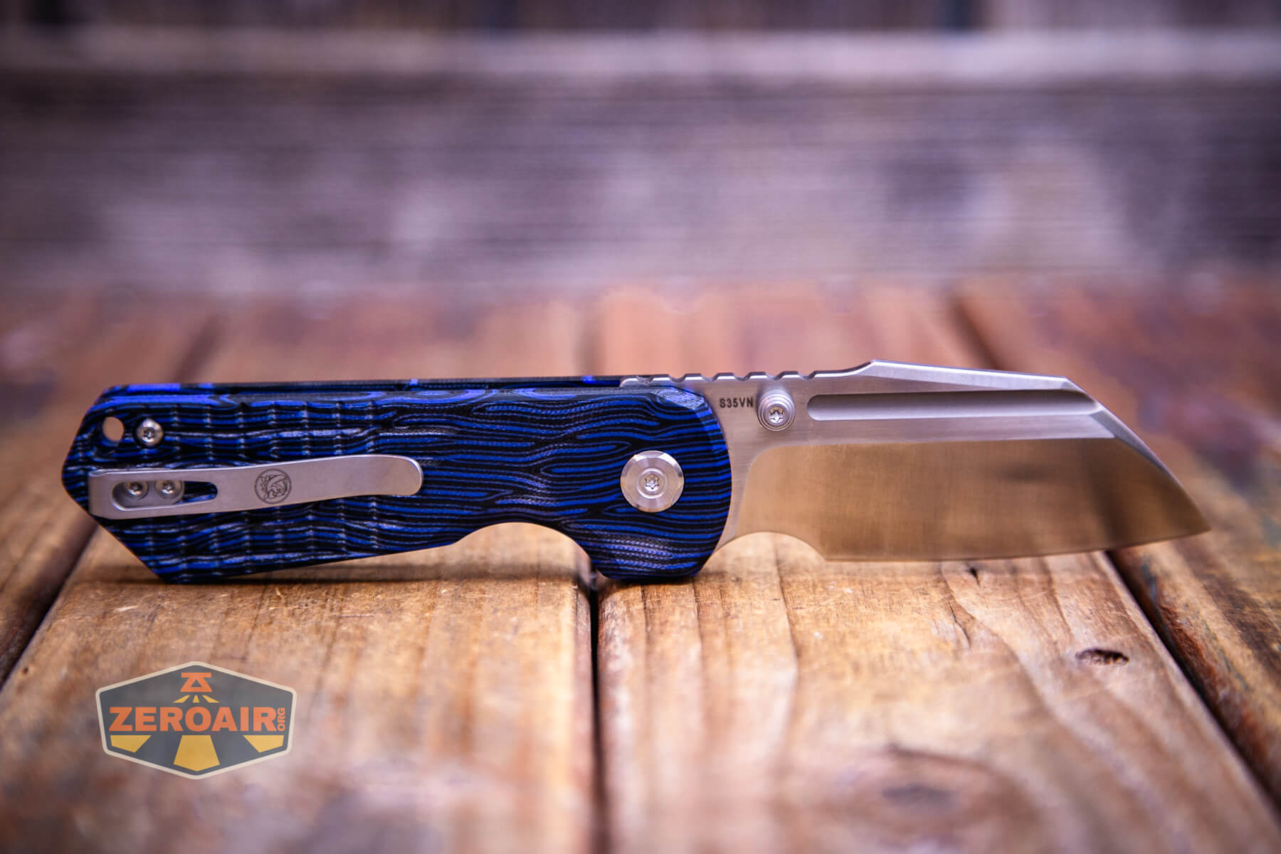A Vosteed Hedgehog Top Liner Lock Knife with a textured blue and black handle and a partially serrated stainless steel blade rests on a wooden surface. The ZeroAir logo is visible in the lower left corner of the image.