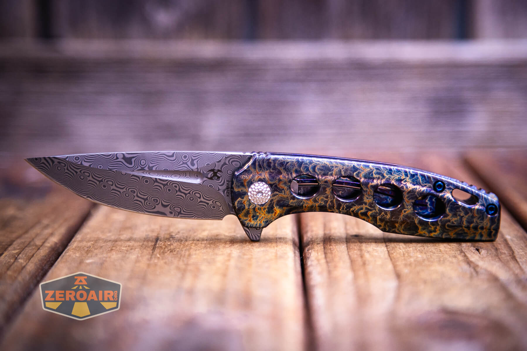 A Kansept Cassowary titanium flipper knife with a patterned blade and a multicolored, textured handle featuring cutout holes rests on a wooden surface. A logo with the text ZEROAIR is visible in the bottom left corner.