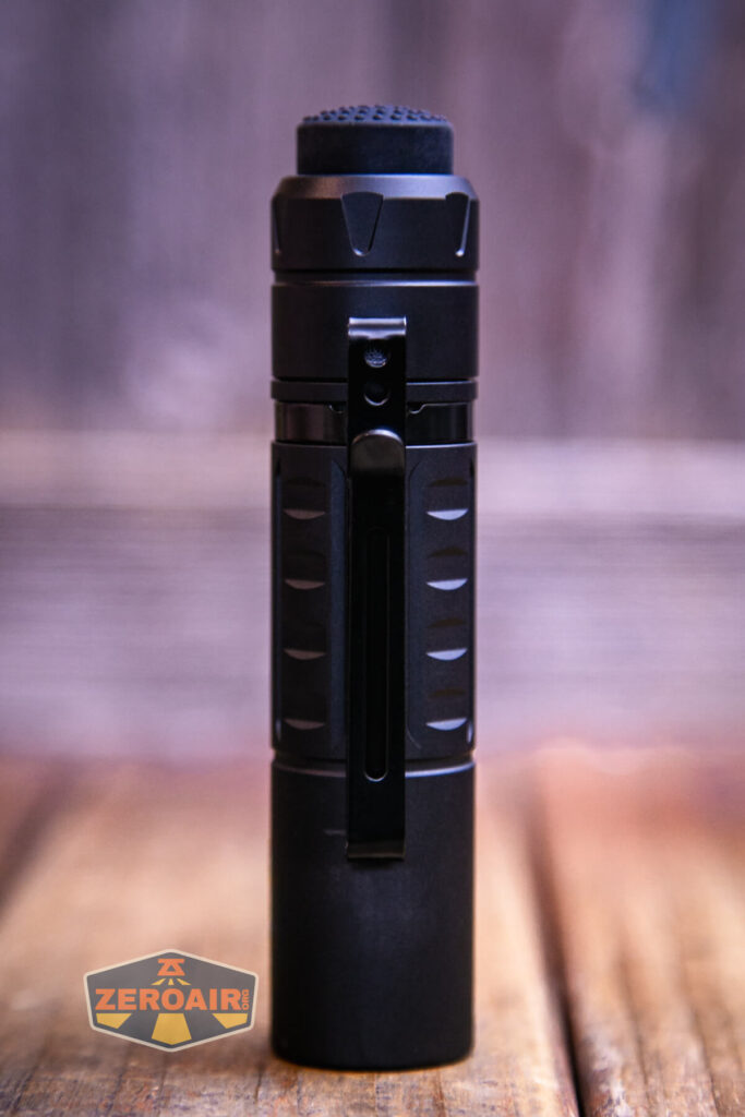 A black cylindrical flashlight with a textured grip and a sturdy pocket clip is standing upright on a wooden surface. The background is blurred, and a ZeroAir watermark is visible in the lower left corner.