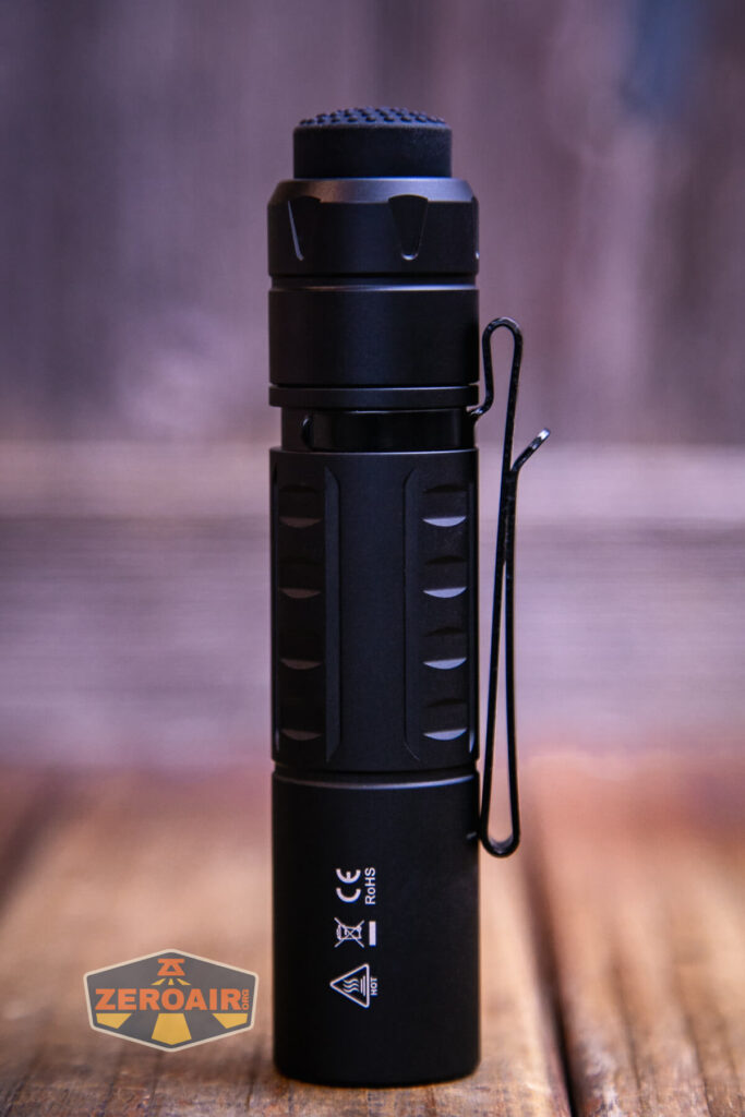 A black, cylindrical flashlight with a textured grip and a side clip stands upright on a wooden surface. The background is blurred, and a small “ZEROAIR” logo is visible in the bottom left corner.