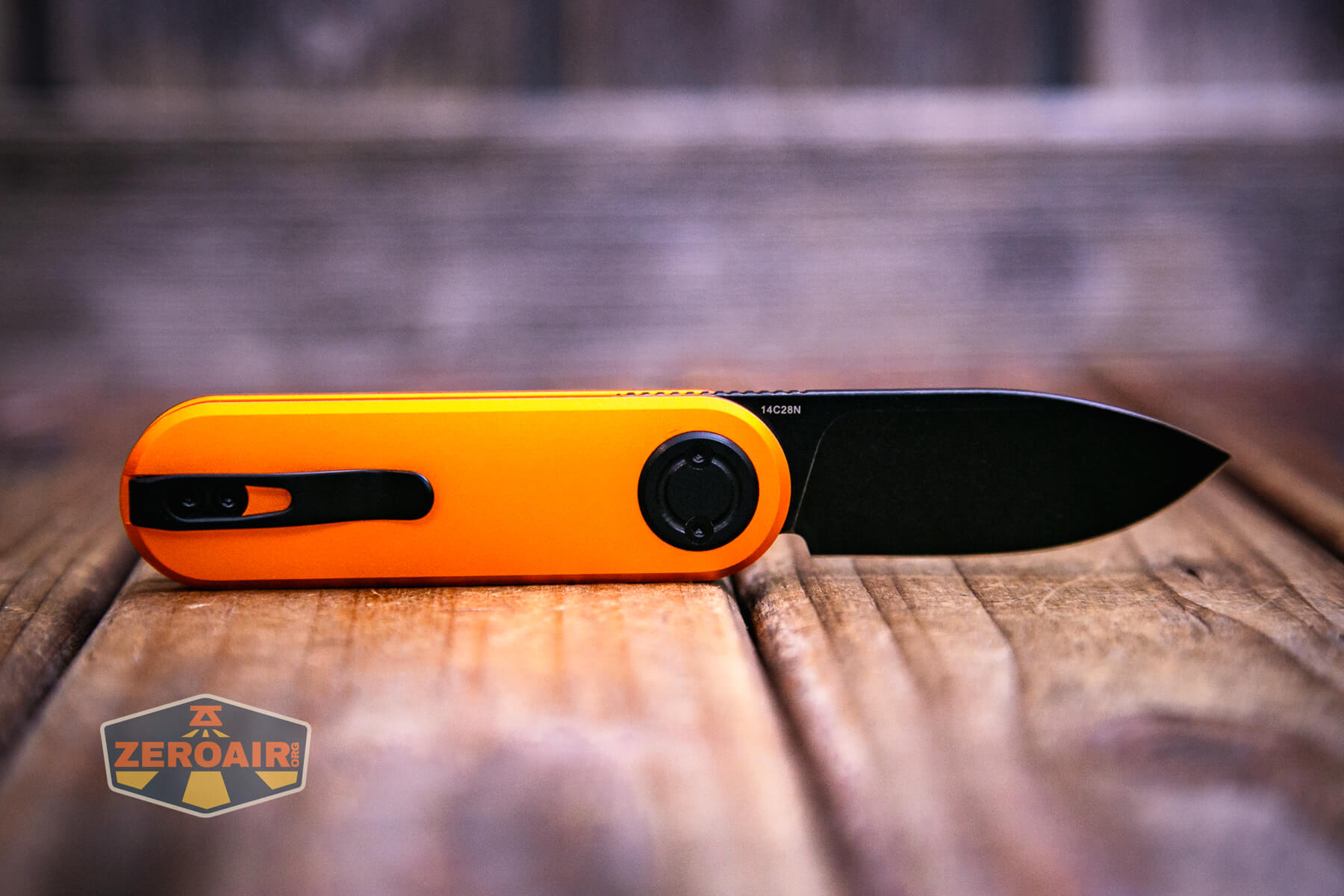 A Vosteed Corgi V knife with an orange handle and black blade rests on a wooden surface. The knife features a handle clip, and the ZeroAir logo is visible in the lower left corner of the image.