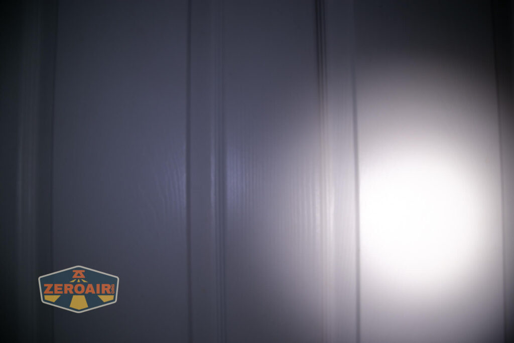 A white door with vertical panels is illuminated by a bright circular flashlight beam on the right side. The ZeroAir logo is visible in the bottom left corner.