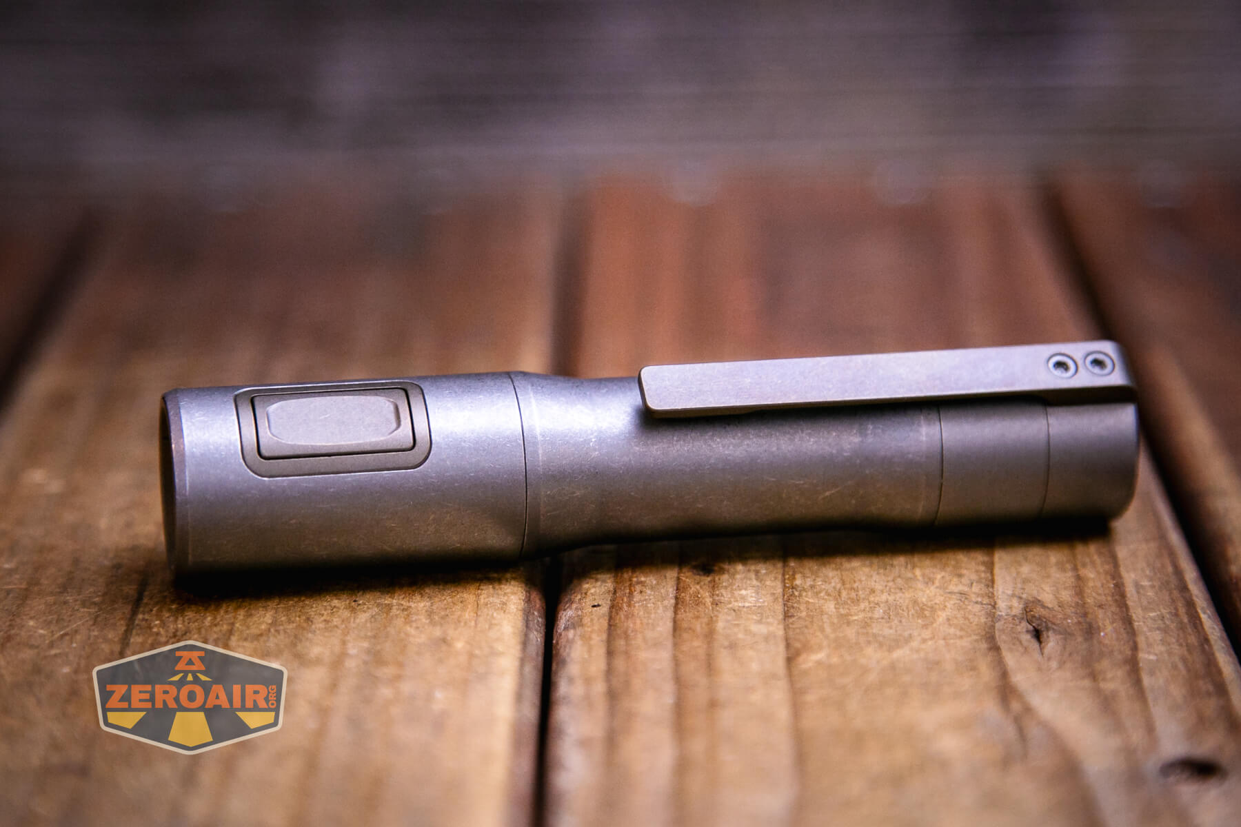 An Exceed Designs Rampant R8 titanium flashlight with a side button and pocket clip lies horizontally on a wooden surface. A small ZeroAir logo appears in the lower left corner of the image.