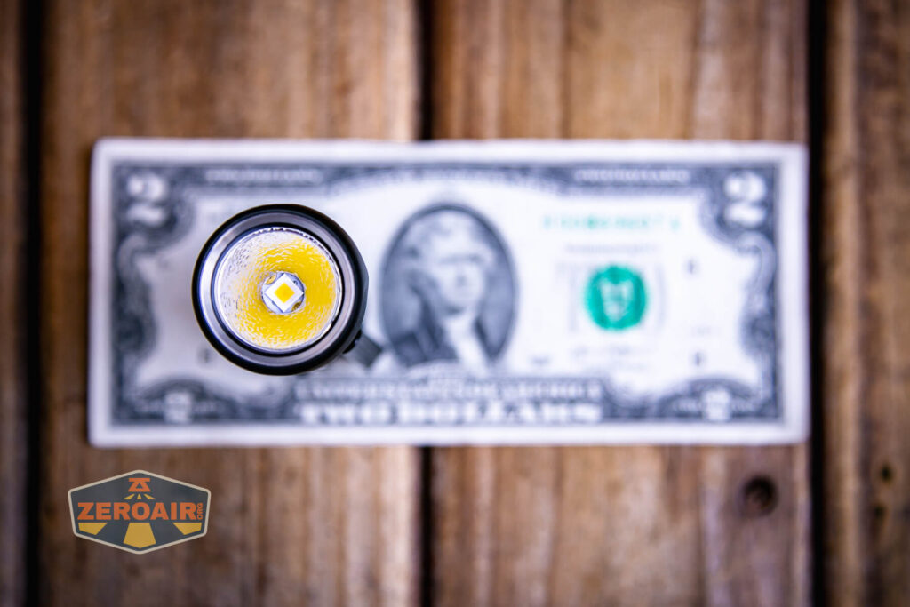 A small flashlight with a lit LED is viewed from above, placed on top of a two-dollar bill on a wooden surface. The ZeroAir logo appears in the bottom left corner of the image.