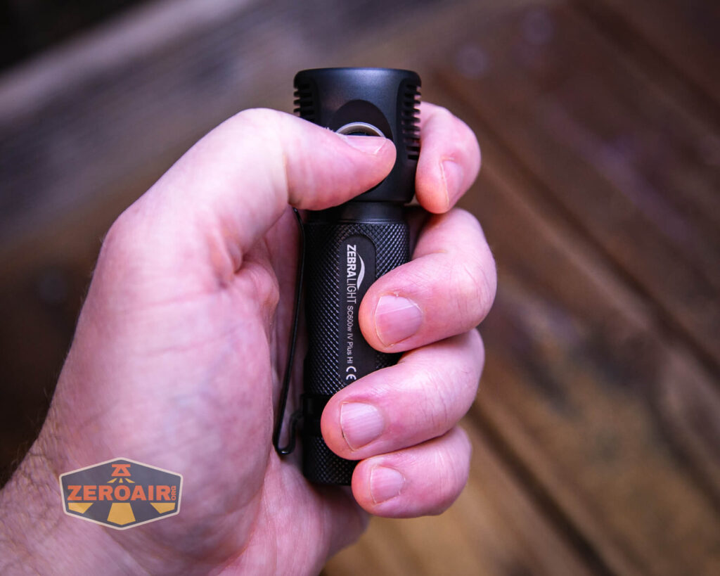 A hand holds a black Zebralight SC600w IV Plus flashlight with a textured grip. The thumb rests on the side button. The background is wooden and slightly out of focus. A ZeroAir logo appears in the lower left corner.