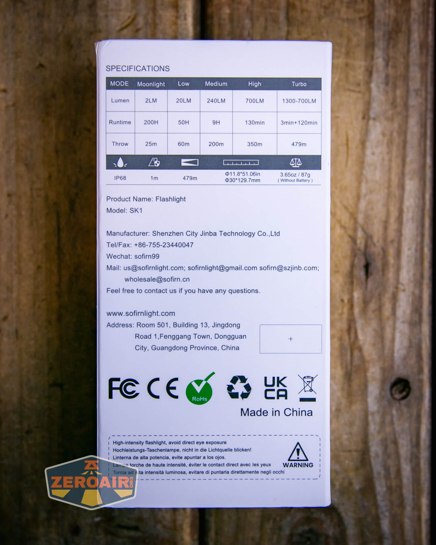 The image shows the back of a white flashlight box with detailed specifications, manufacturer information, website, and certification logos, set on a wooden surface with a ZeroAir sticker in the bottom left corner.