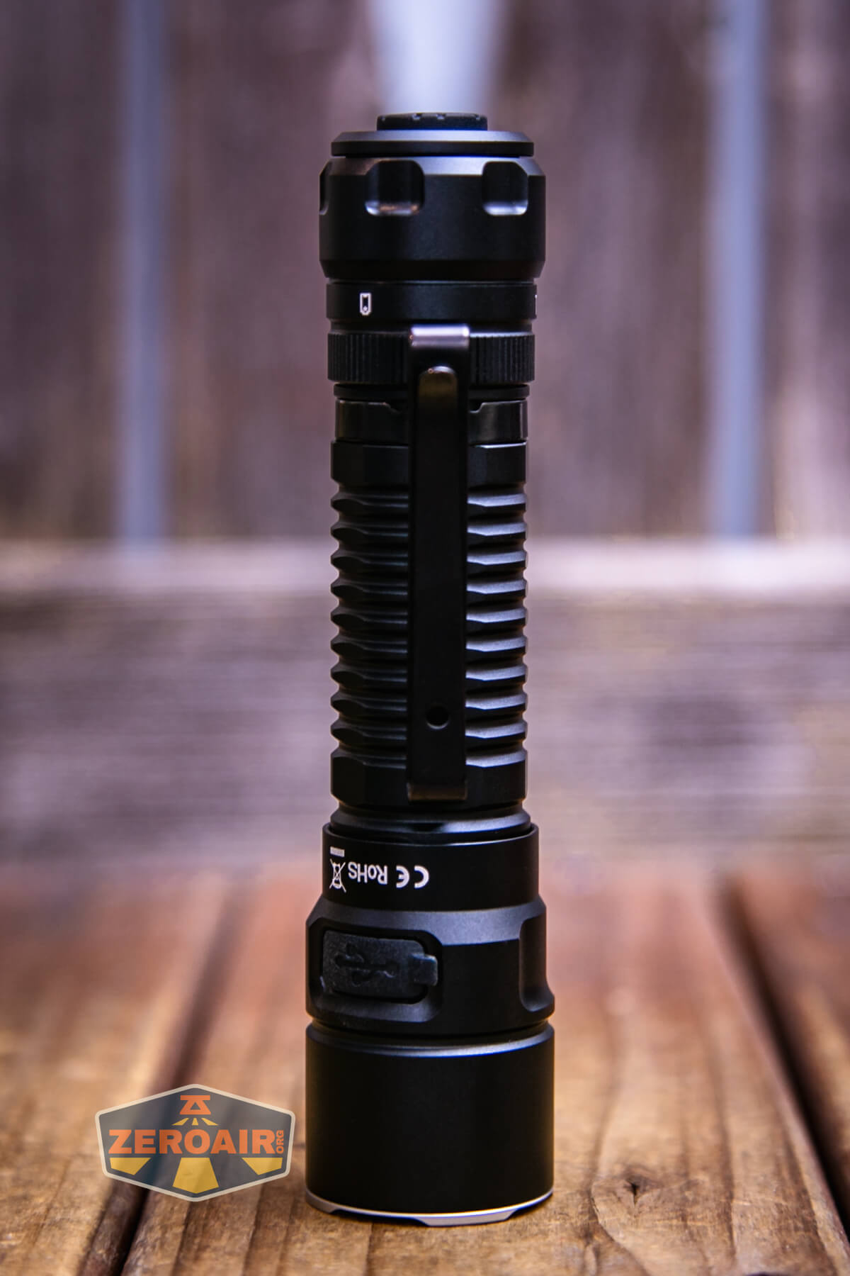 A black, cylindrical tactical flashlight stands upright on a wooden surface with a blurred wooden background. A ZEROAIR logo is visible in the bottom left corner of the image.