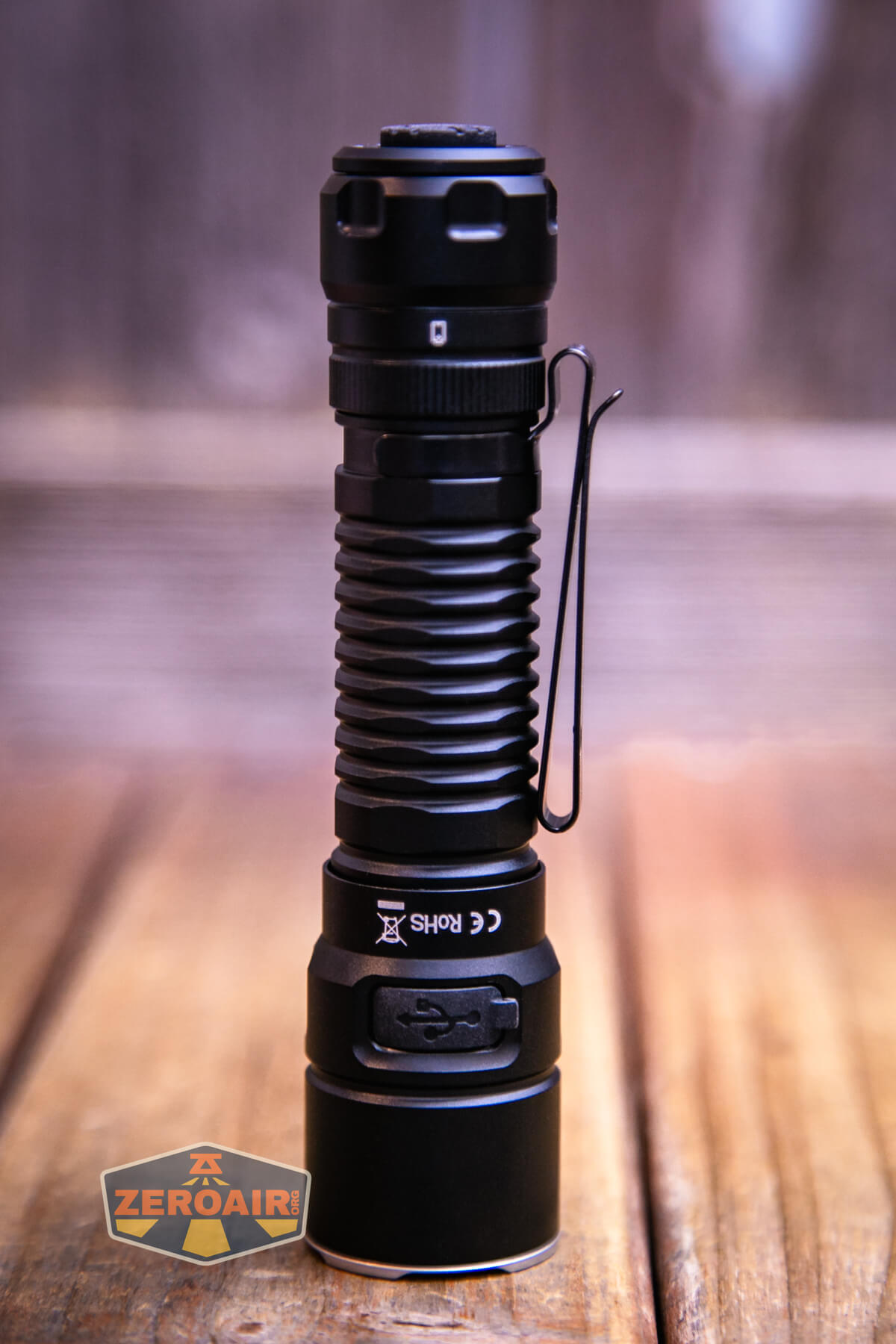 A black, cylindrical flashlight with ribbed grip and metal pocket clip stands upright on a wooden surface. The background is blurred. The ZeroAir logo is in the lower left corner.