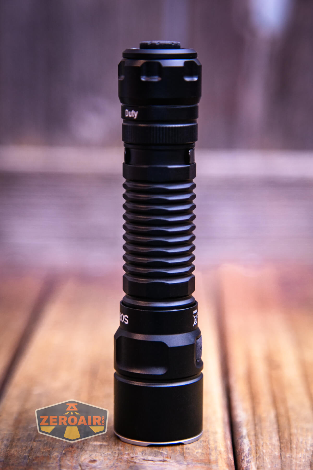 A black tactical flashlight is standing upright on a wooden surface. The background is blurred, and a ZeroAir logo is visible in the bottom left corner of the image.