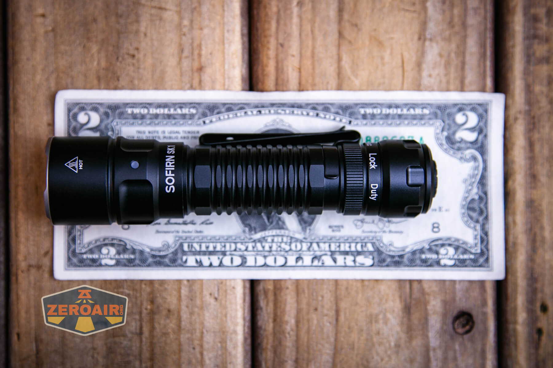 A black Sofirn flashlight is placed horizontally on top of a U.S. two-dollar bill, which is on a wooden surface. A ZeroAir logo is visible in the bottom left corner of the image.