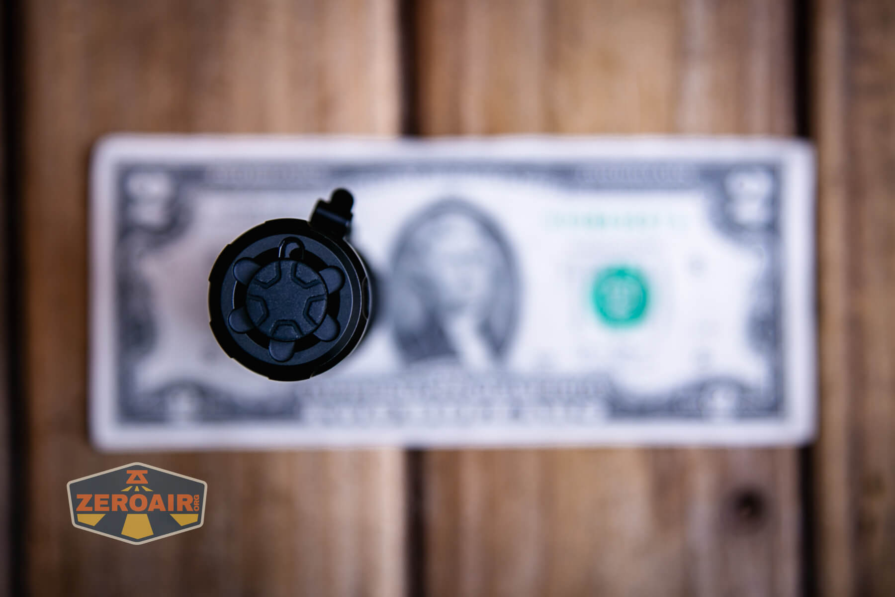 A Sofirn SK1 tactical flashlight, black and cylindrical, is viewed from above and centered over a U.S. one-dollar bill on a wooden surface. The image includes a ZEROAIR! logo in the bottom left corner.
