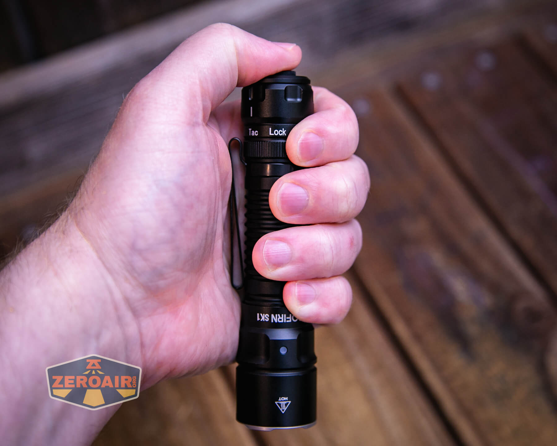 A close-up of a persons hand holding a black Olight Seeker flashlight, with the thumb near the lock switch. The background consists of a wooden surface, and a ZeroAir logo is visible in the bottom left corner.