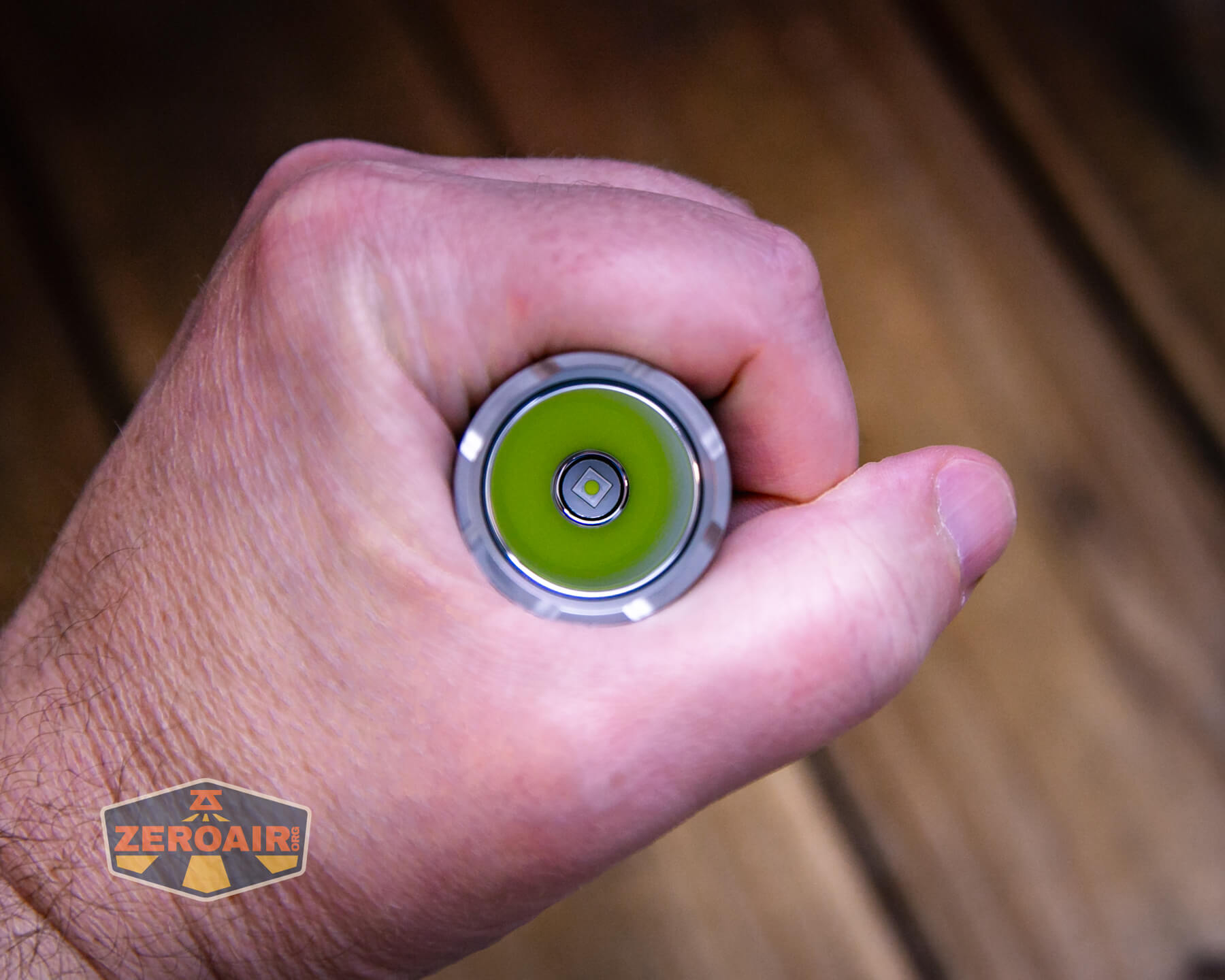 A hand grips a flashlight with the lens facing the camera, showing a green circuit and LED at the center; wooden floorboards are visible in the background and a ZeroAir logo appears in the bottom left corner.