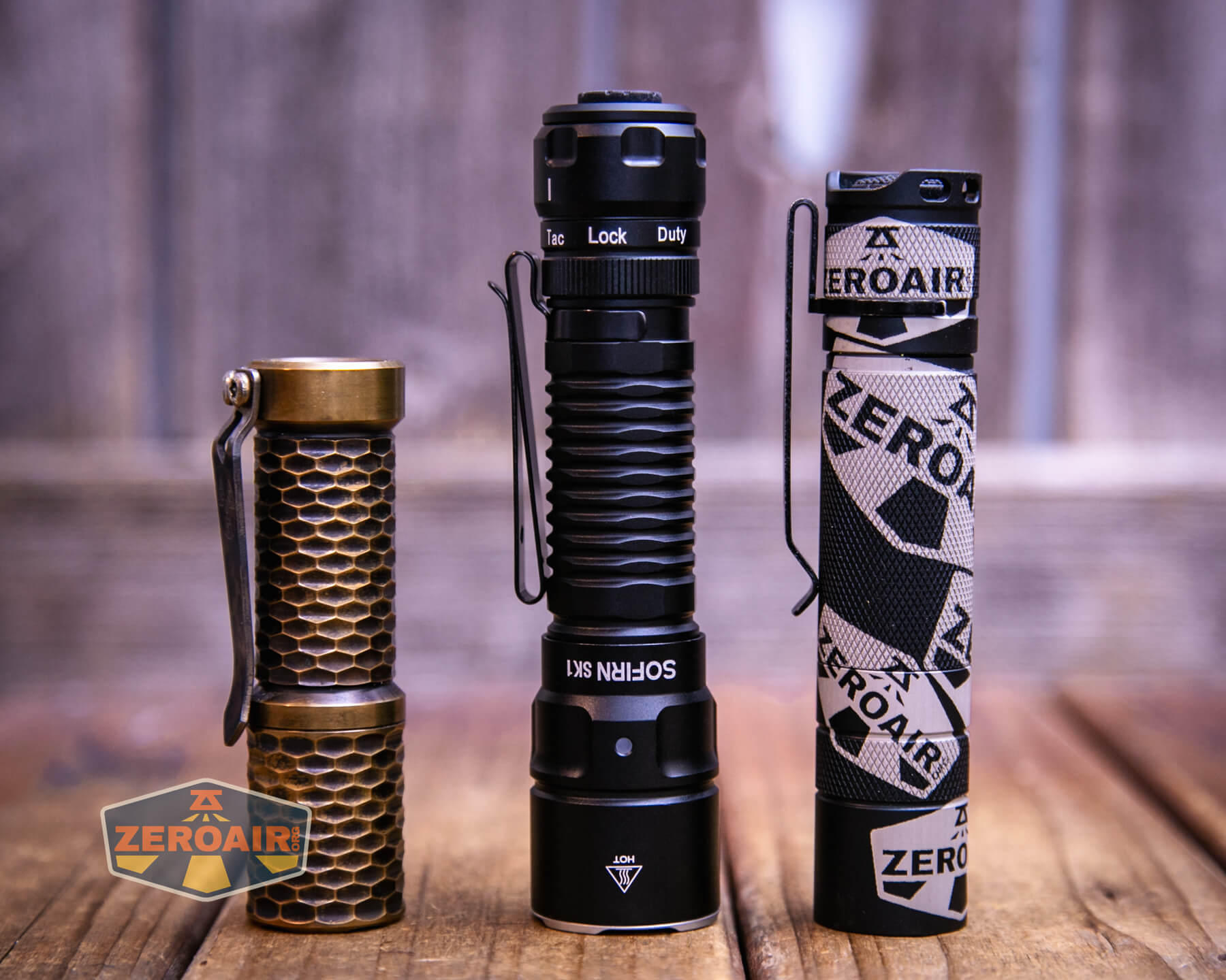 Three flashlights stand upright side by side on a wooden surface. The left one has a honeycomb pattern and a bronze finish, the middle is black and labeled “Sofirn S21,” and the right is black with a printed ZeroAir cover.