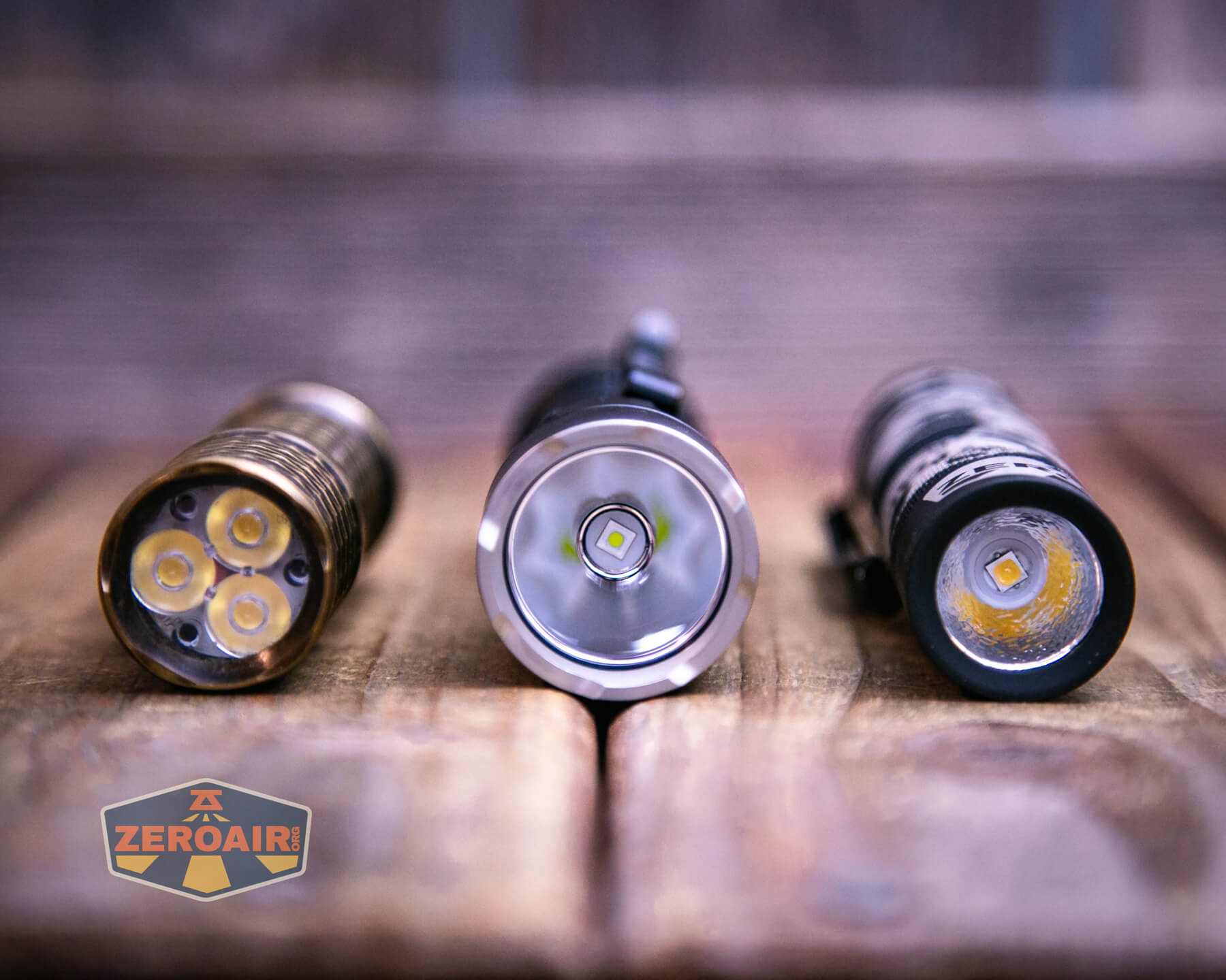 Three flashlights are positioned upright on a wooden surface. The image shows their LED emitters facing the camera. A ZeroAir logo is visible in the bottom left corner. The background is slightly blurred.