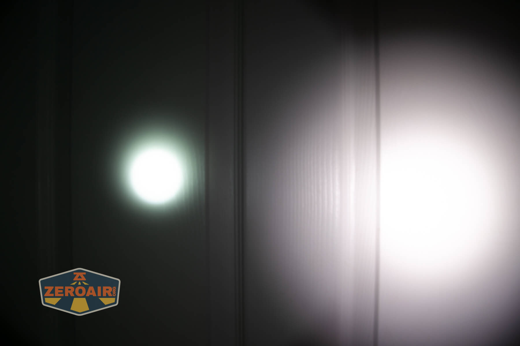 A dark room with two flashlight beams, one narrow and greenish on the left, and one wider and white on the right, shining on a paneled wall. A ZEROAIR logo appears in the lower left corner.