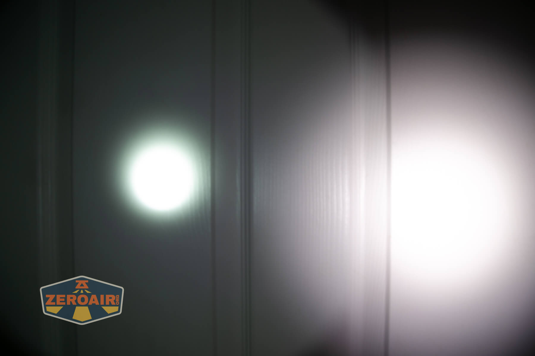 Two flashlight beams of different brightness and sizes are projected onto a white paneled wall; the left beam is smaller and more focused, while the right beam is larger and diffused. A ZeroAir logo is in the bottom left corner.