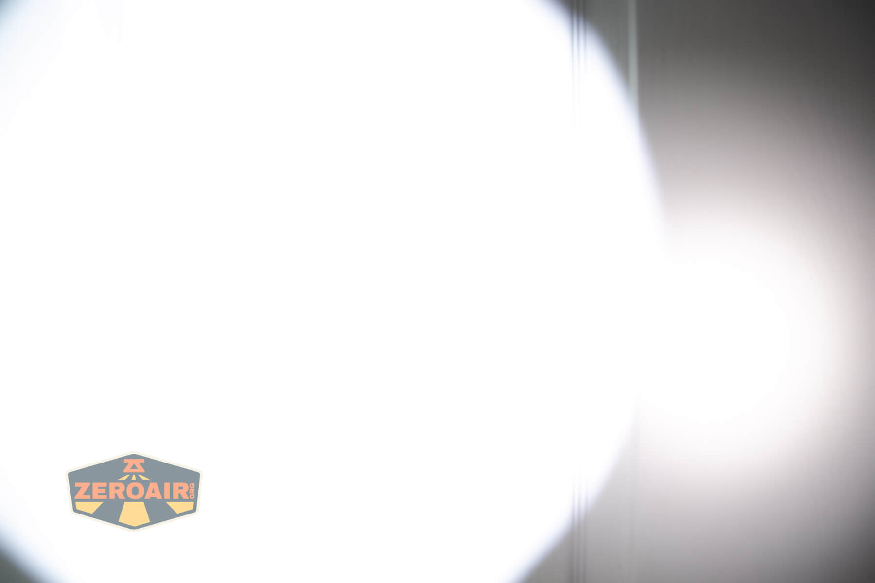 A bright white circular light beam is cast on a neutral wall; the light fades out toward the edges. In the bottom left corner, there is a Zeroair logo with a black and yellow design.