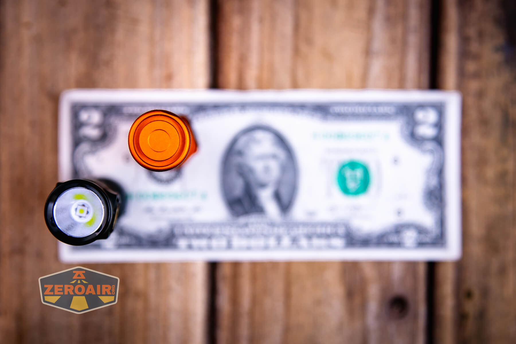 A close-up view of the tops of two cylindrical objects, one orange and one black, placed on top of a two-dollar bill on a wooden surface. A logo reading ZEROAIR appears in the lower left corner.