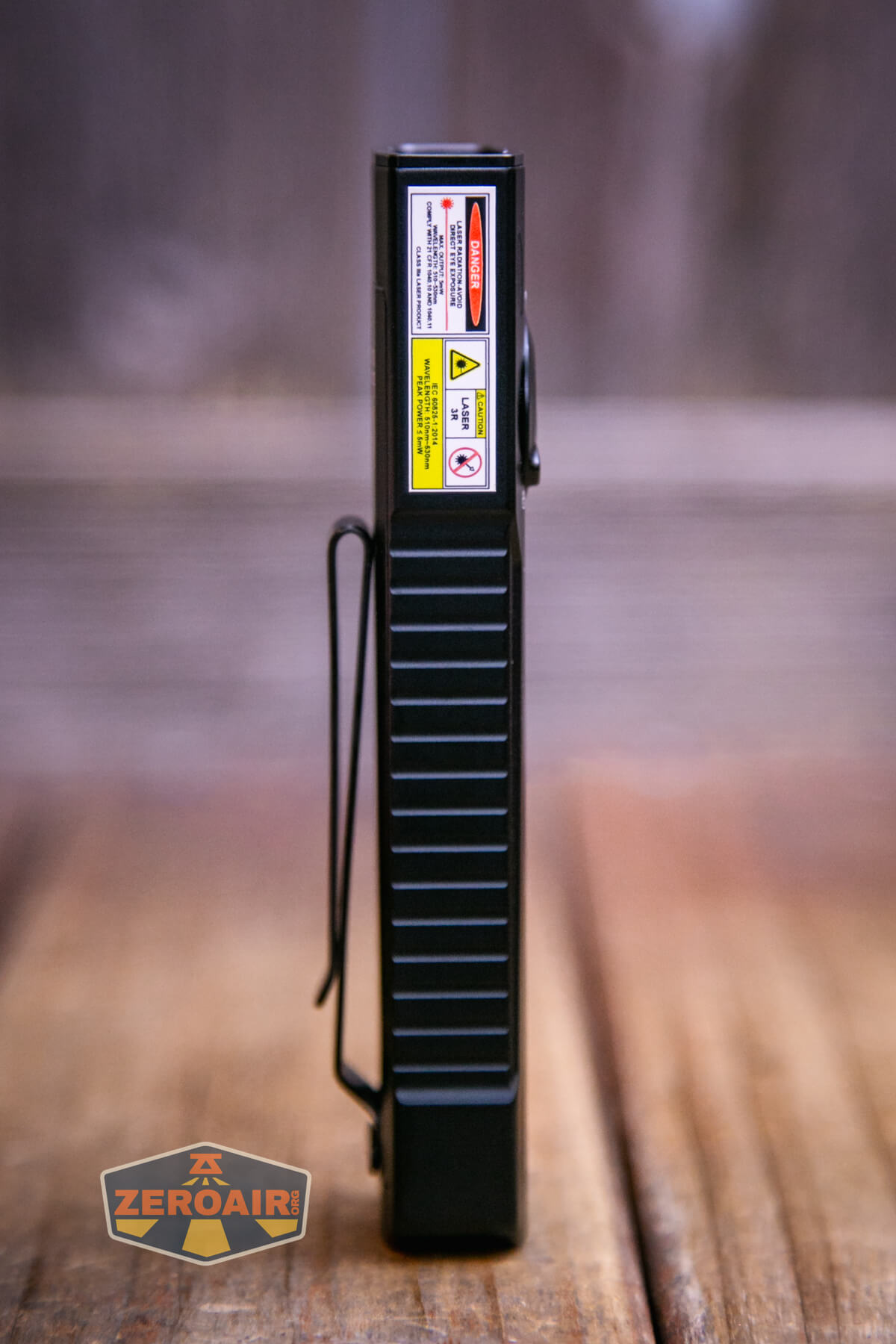 A rectangular black device stands upright on a wooden surface with a metal clip on the side and caution labels on its upper edge. The background is out of focus. A ZeroAir logo is in the bottom left corner.