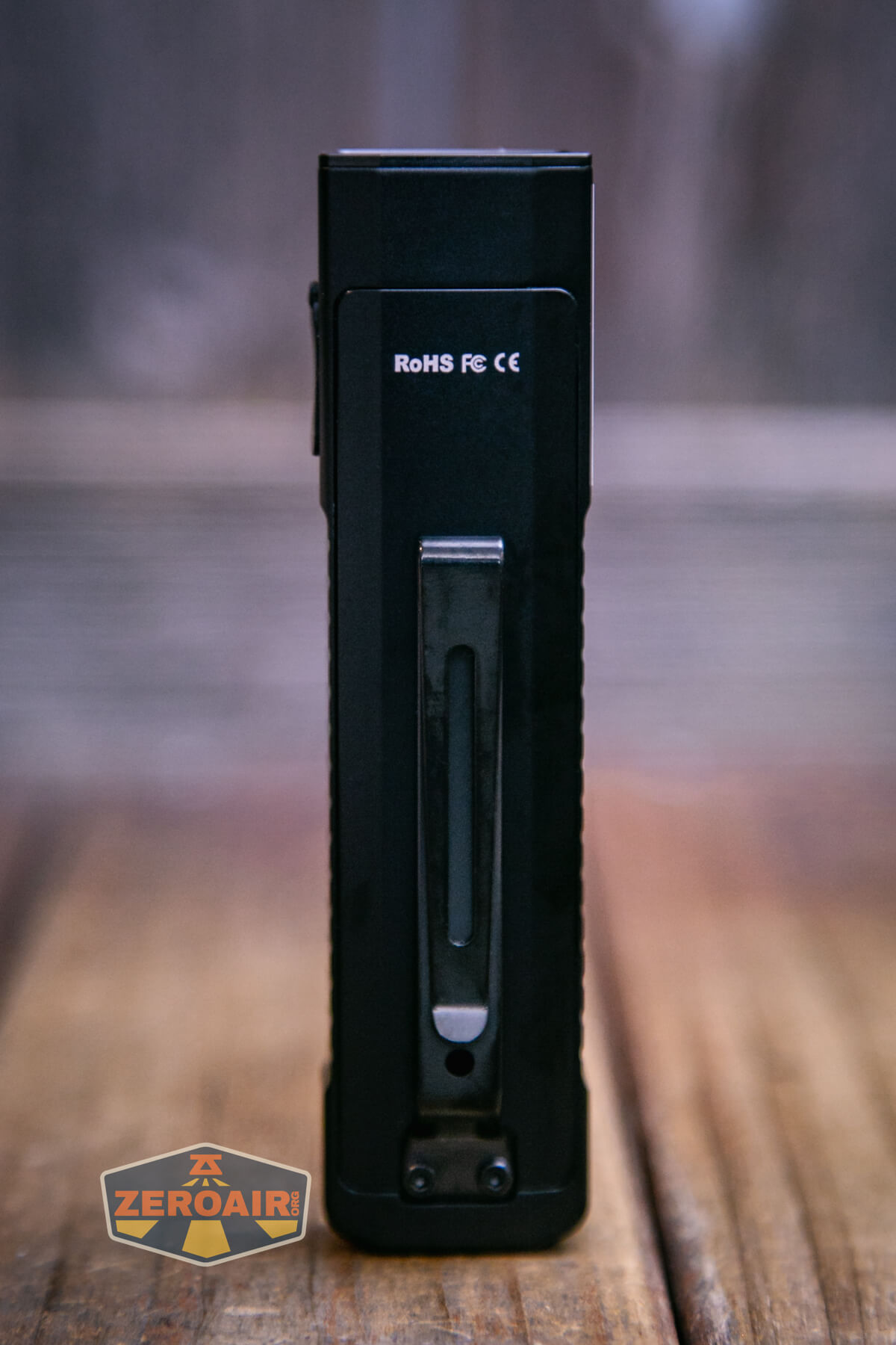 A black rectangular electronic device stands upright on a wooden surface, featuring a metal clip on its front and white certification markings near the top. A ZeroAir logo is visible in the lower left corner.