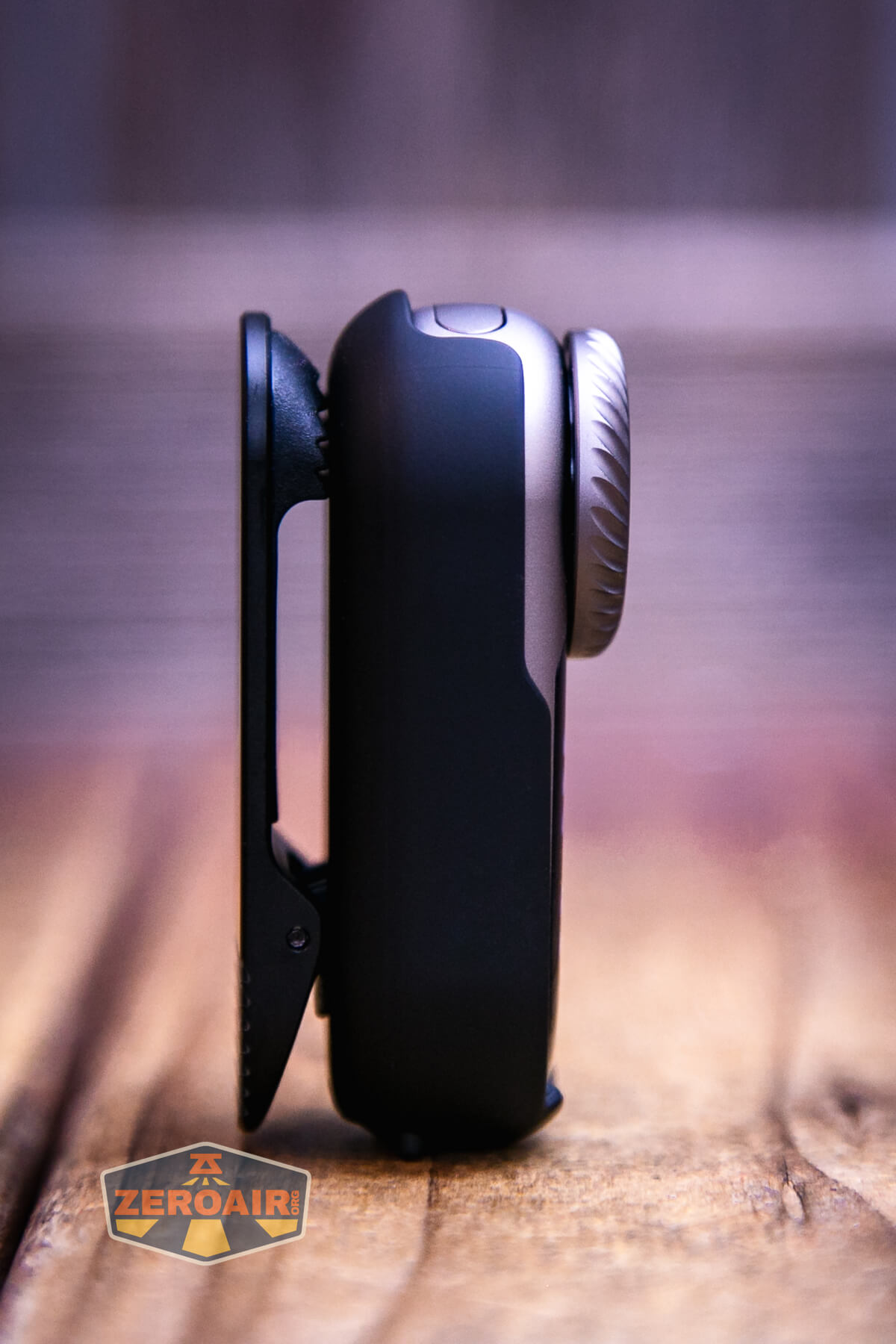A close-up side view of a small black device with a ridged circular dial and clip, standing vertically on a wooden surface. The ZeroAir logo is visible in the bottom left corner.