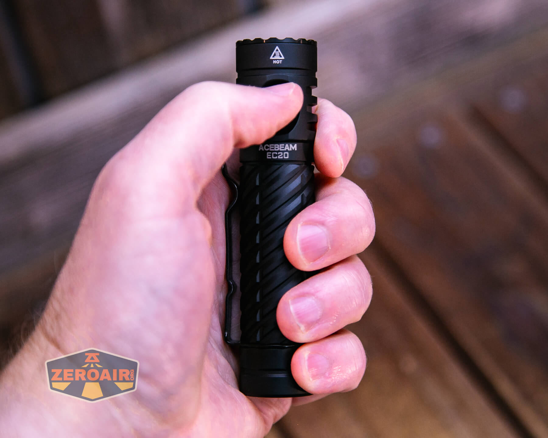 A person holding a black Acebeam EC20 flashlight in their right hand, with a wooden surface in the background and a small ZeroAir logo in the lower left corner.