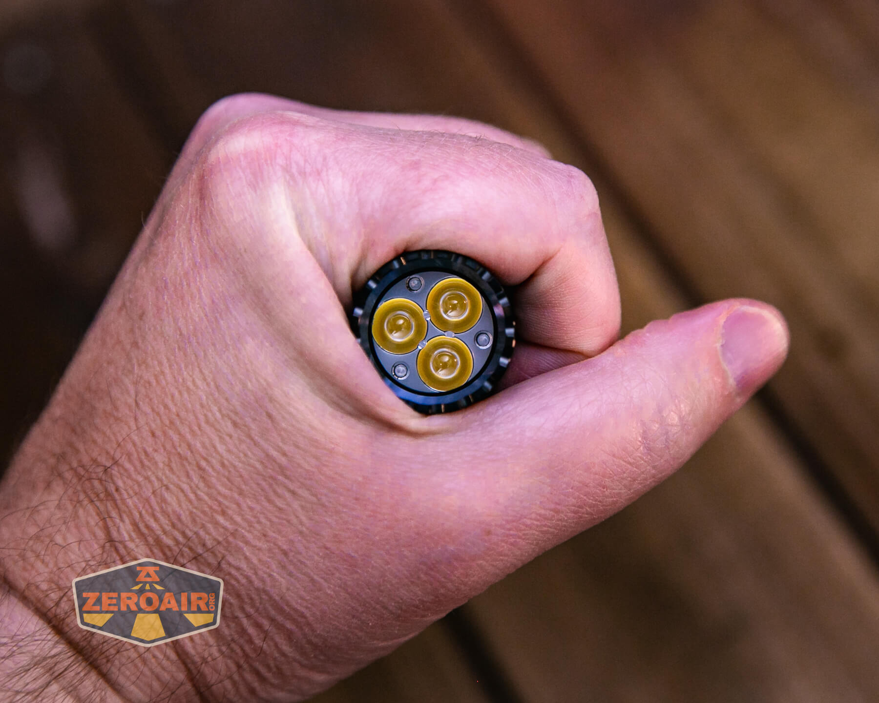 A hand holding a flashlight with three visible LED bulbs, viewed from above. The flashlight is gripped tightly, and a “ZeroAir” logo is visible in the lower left corner. The background is blurred brown wood.