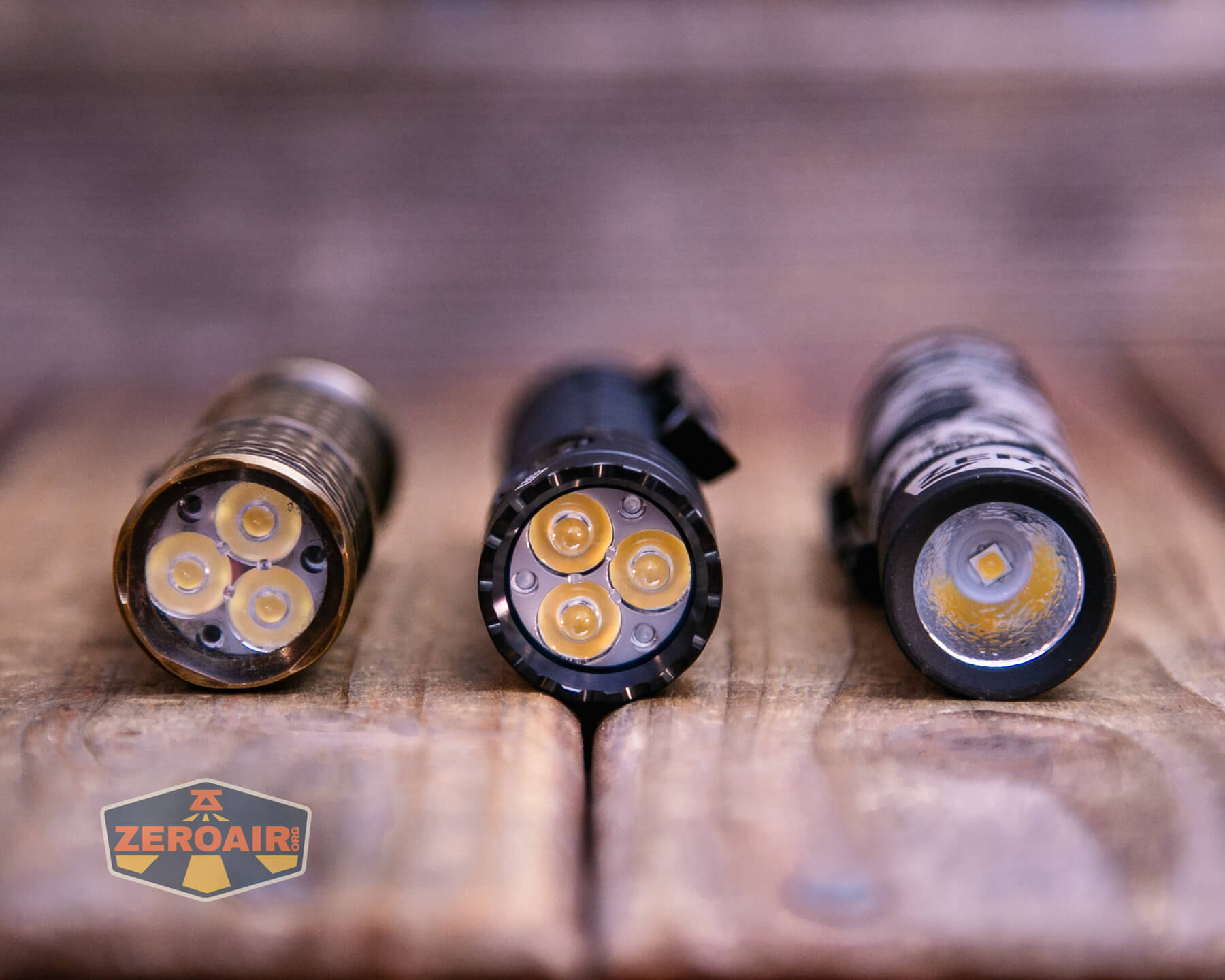 Three flashlights are placed on a wooden surface, shown from the front to display their LED bulbs. Each flashlight has a different arrangement of LEDs. A ZeroAir logo is visible in the lower-left corner.