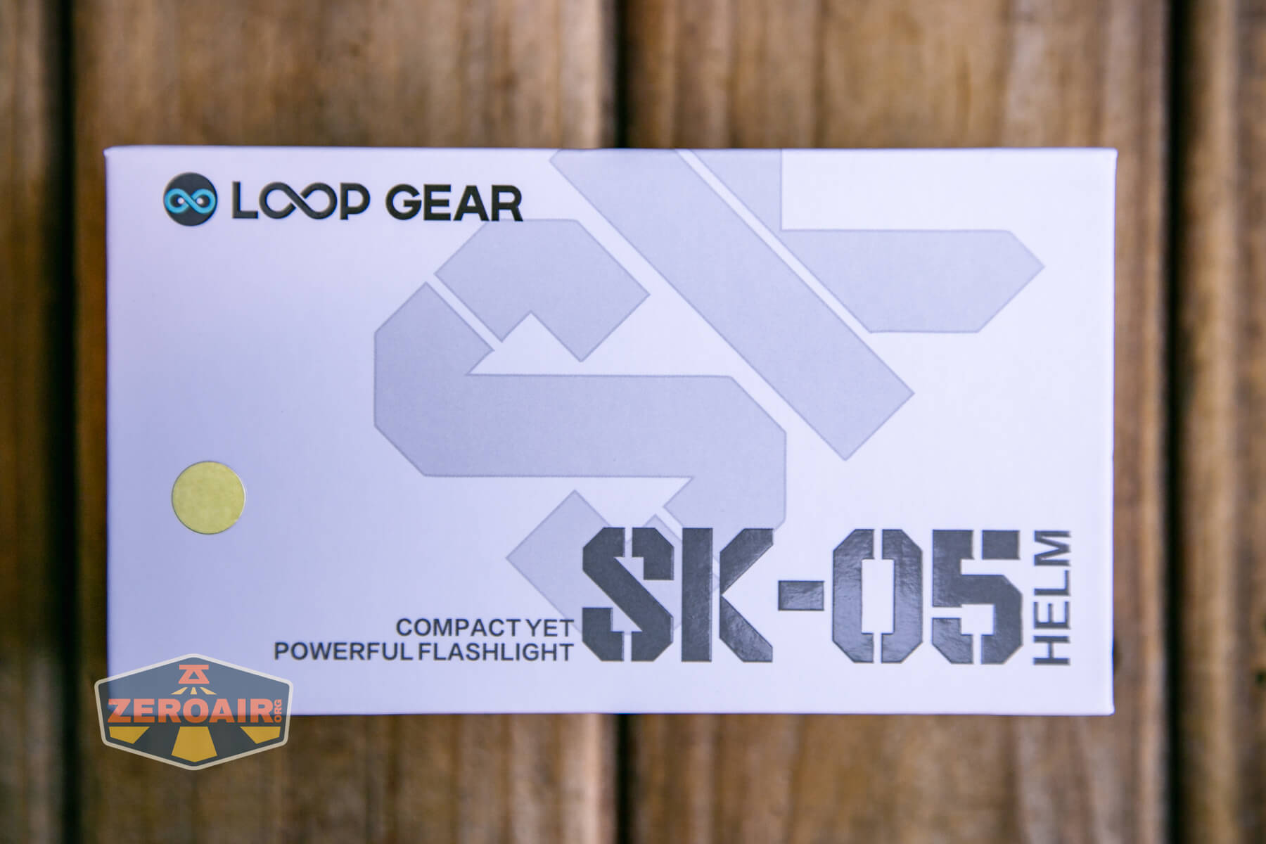 A white box with the text “LOOP GEAR SK-05 HELM Compact Yet Powerful Flashlight” printed on it, placed on a wooden surface. The ZeroAir logo is in the bottom left corner of the image.