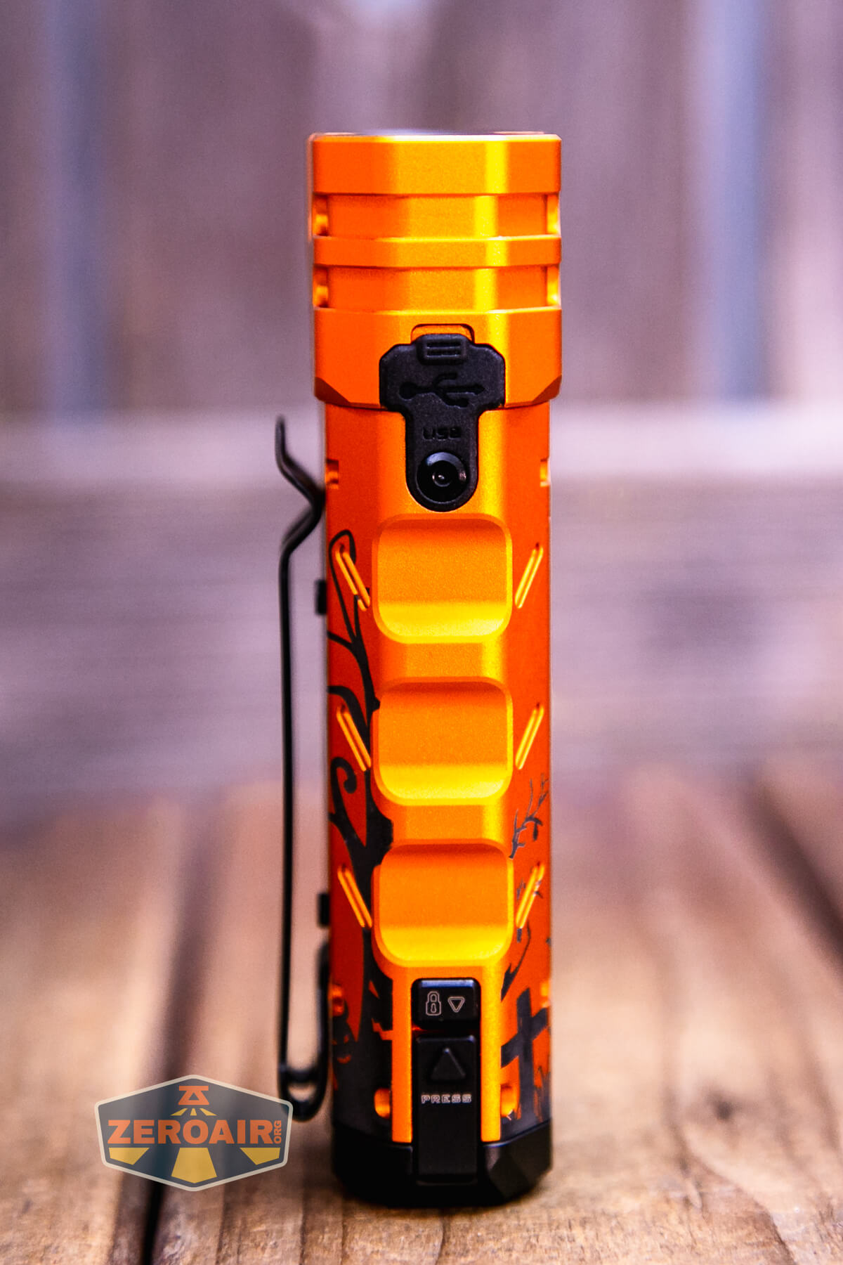 An orange flashlight with a geometric grip pattern and black decorative markings standing upright on a wooden surface. A metal clip is attached to the side, and a USB port cover is visible near the top.