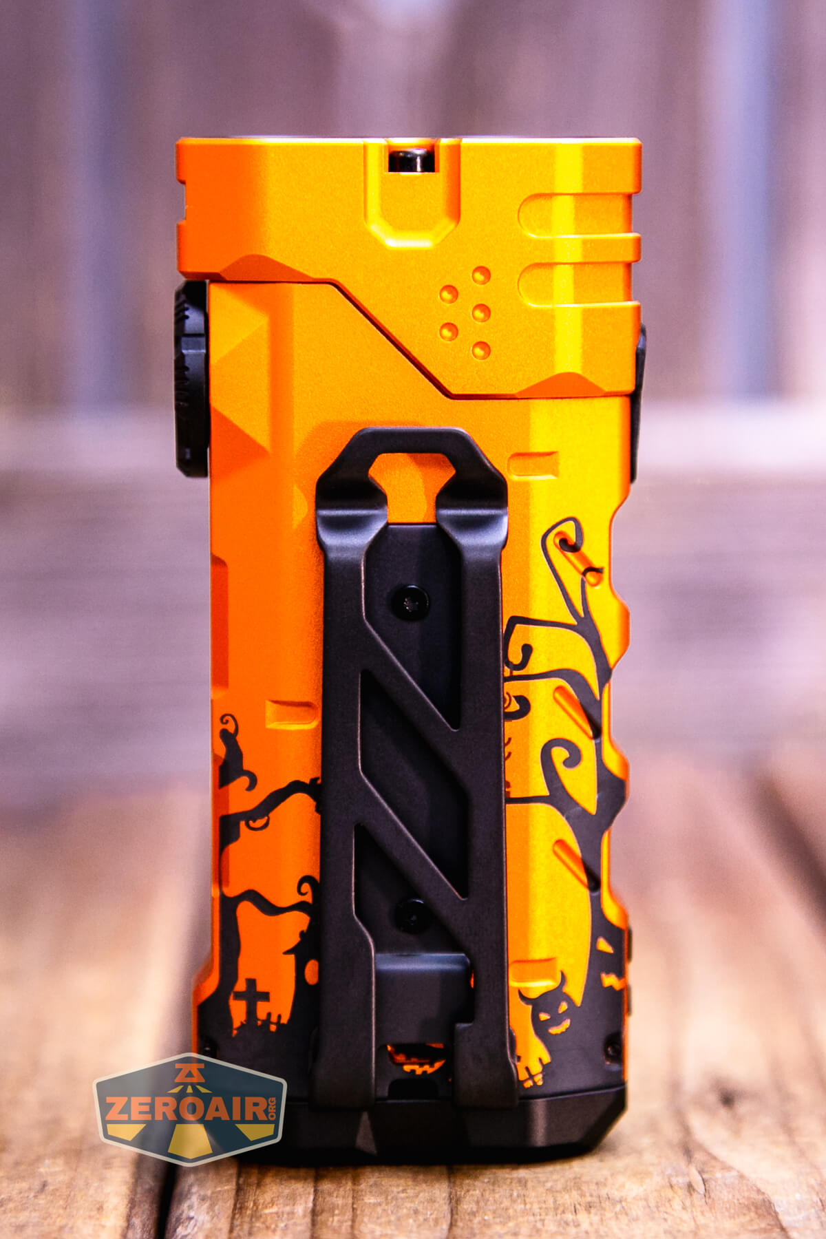 A bright orange rectangular flashlight with black cat and pumpkin silhouettes in a Halloween theme, featuring a black clip. It is standing upright on a wooden surface with a blurred background.