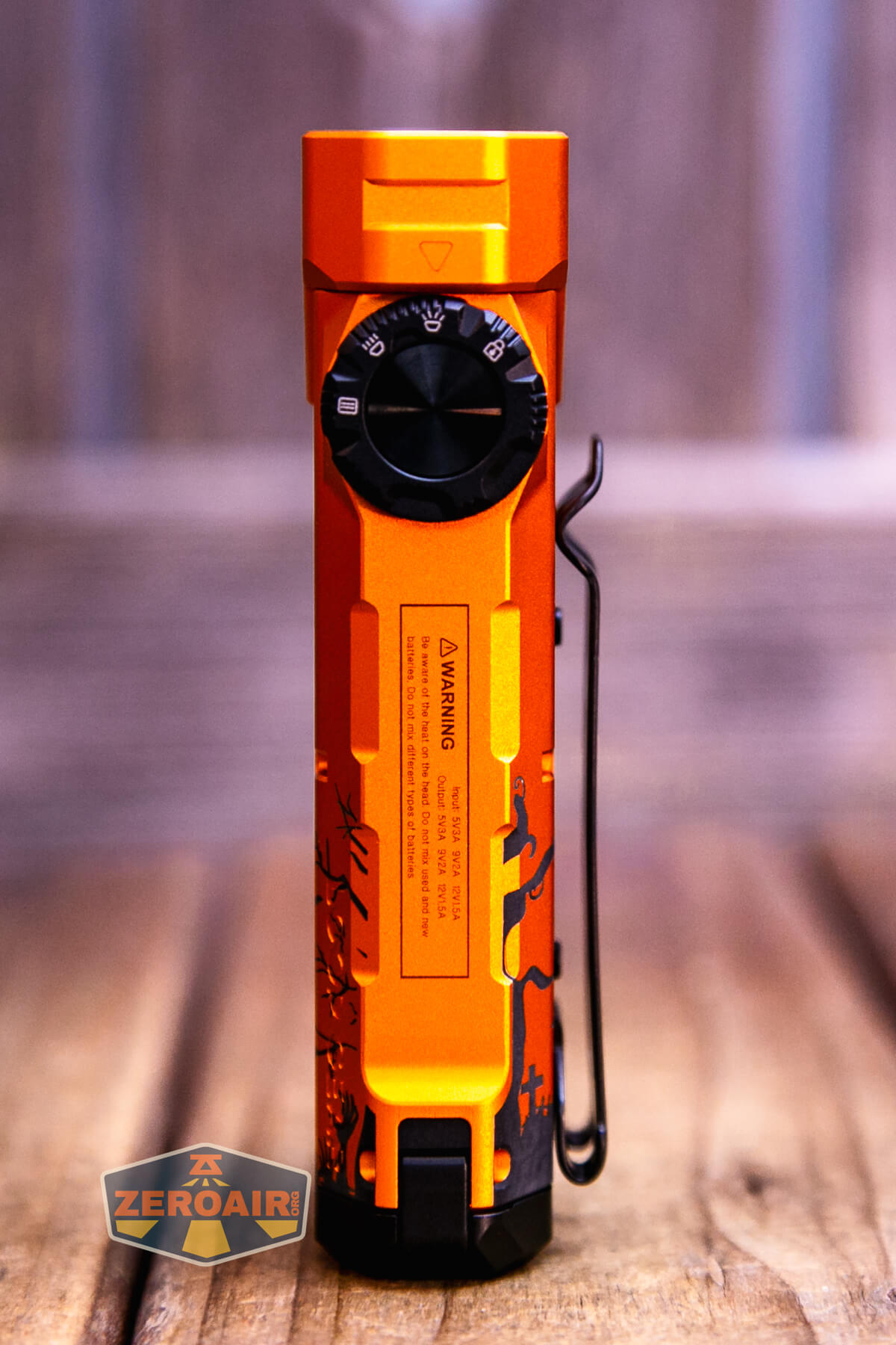 An orange flashlight with a black side switch, a side clip, and warning label, standing upright on a wooden surface with a blurred wood background. The flashlight features a pattern on its lower section.