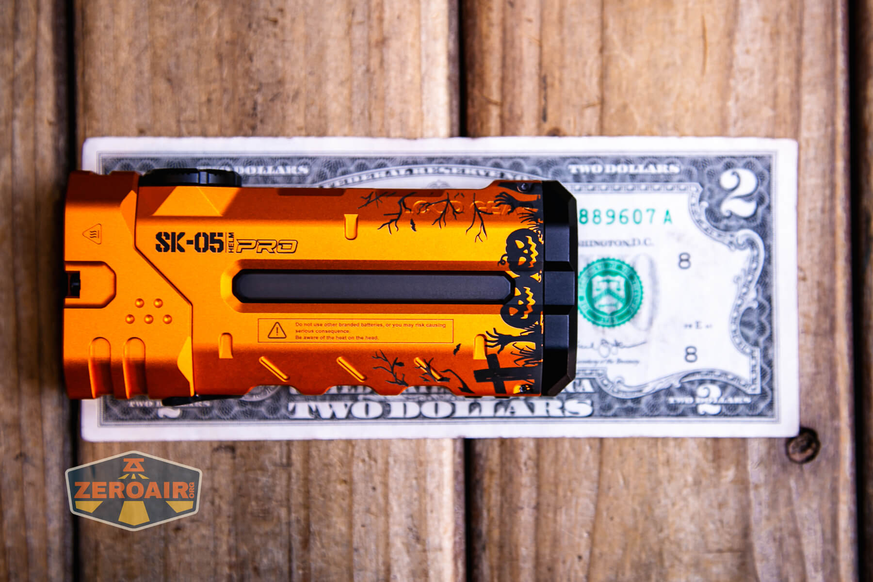 An orange SK-051 PRO flashlight lies on a two-dollar bill atop a wooden surface. A ZeroAir logo sticker is placed in the bottom left corner of the image.