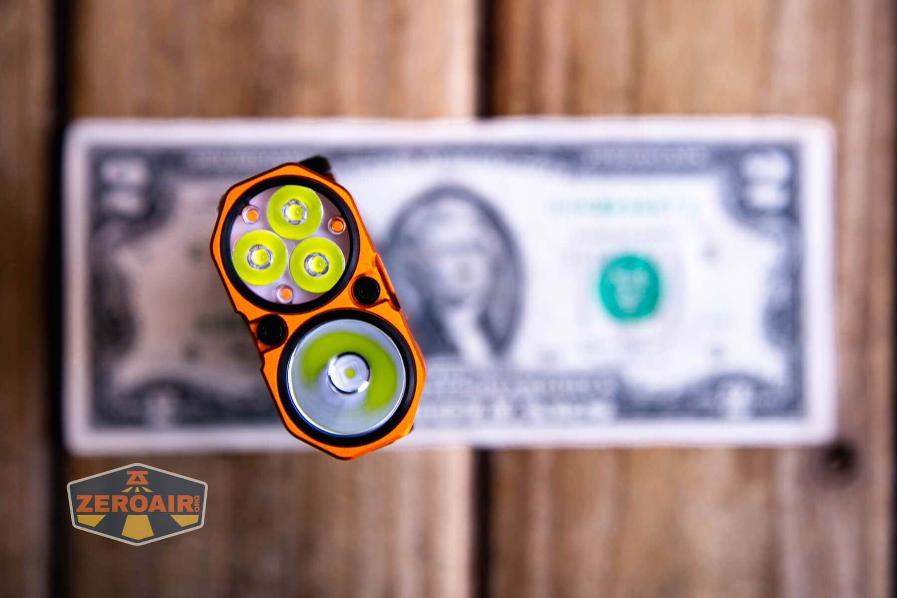 An orange flashlight with four LED bulbs is shown from above, positioned in front of a blurry U.S. one dollar bill on a wooden surface. A ZeroAir sticker is visible in the lower left corner.