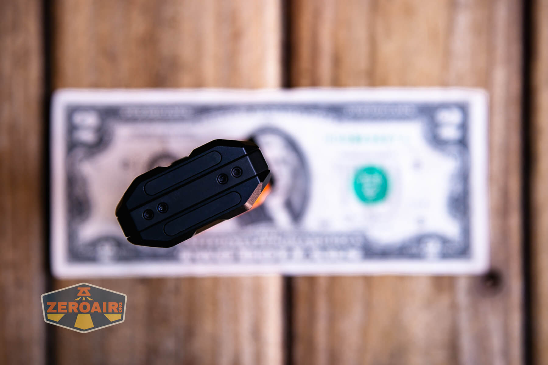 A black rectangular object is placed upright on top of a blurred two-dollar bill on a wooden surface. The image includes a ZeroAir logo in the lower left corner.