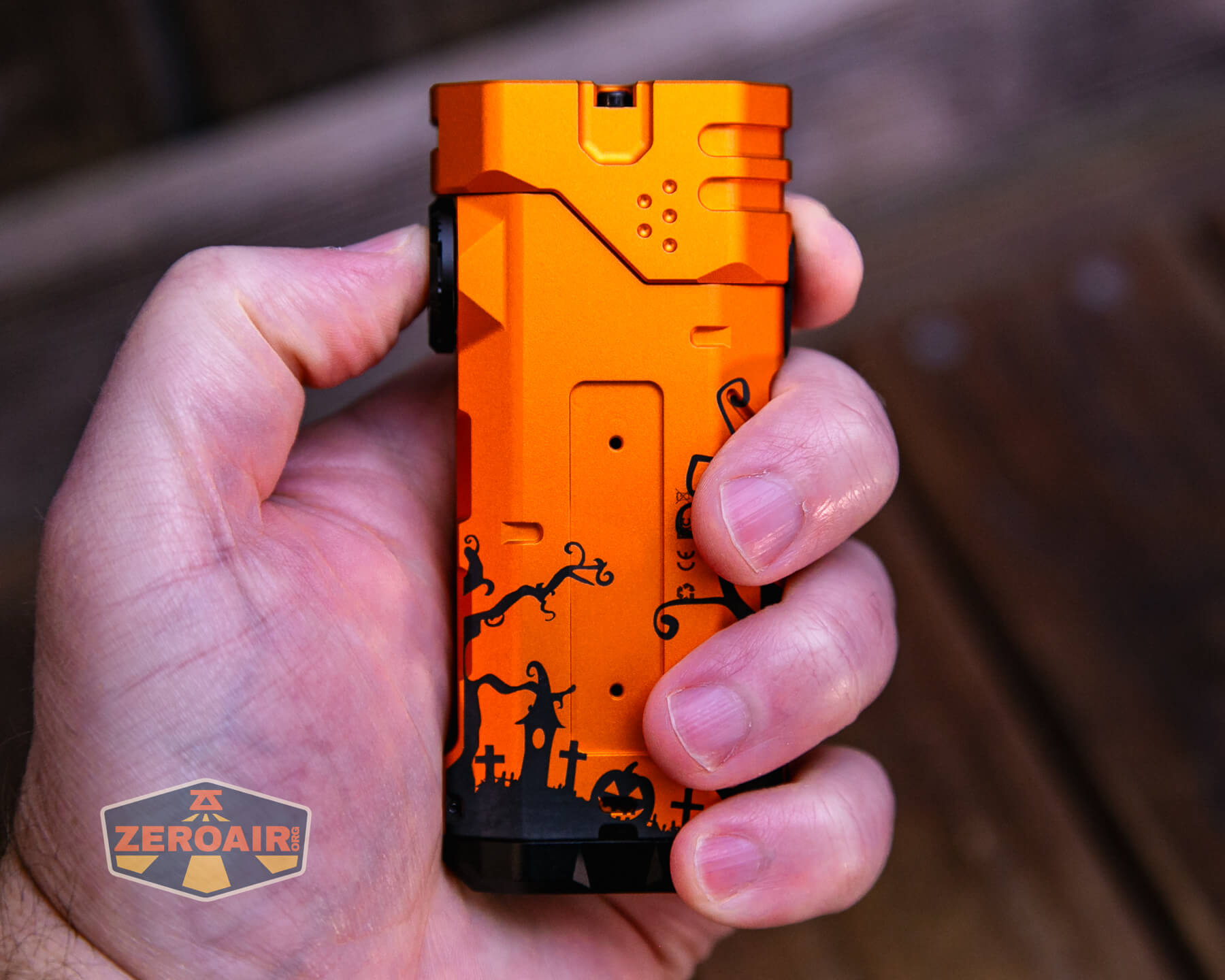 A hand holds an orange rectangular flashlight with a black Halloween-themed design, including pumpkins and twisted trees. The flashlight has a button on the side, and a ZeroAir logo is visible in the bottom left corner.