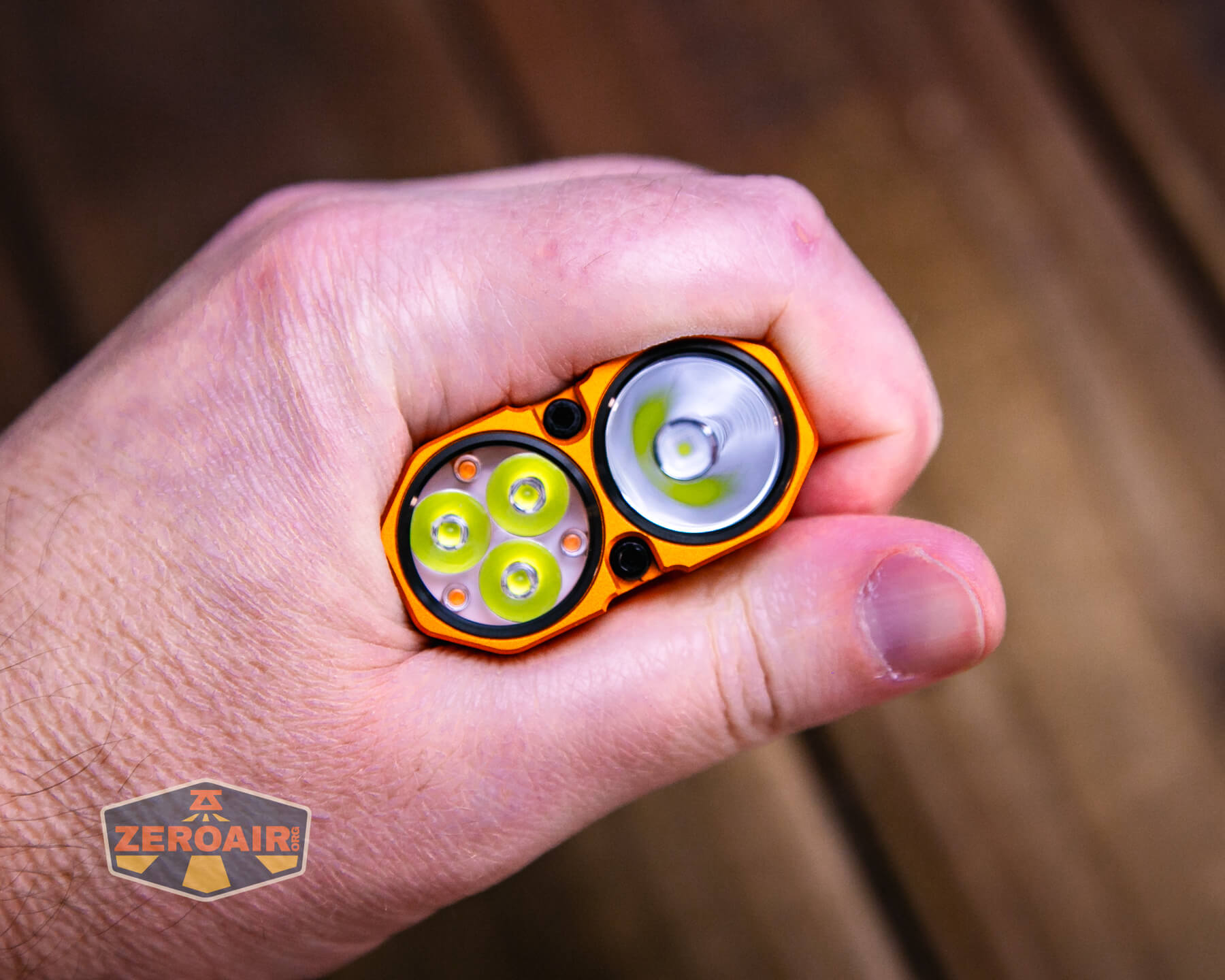 A hand holding an orange flashlight with two heads: one featuring four small LED bulbs and the other with a single larger LED bulb. The background is a wooden surface, and a ZeroAir logo is visible in the corner.