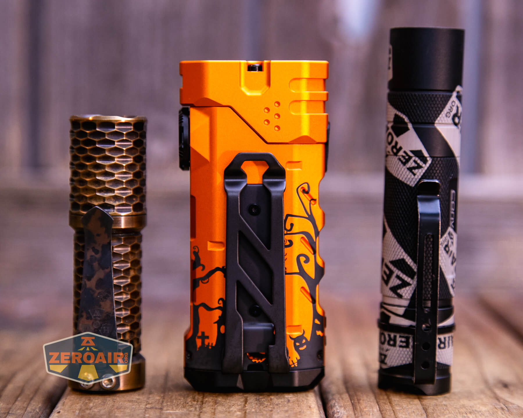 Three flashlights stand upright on a wooden surface. The left flashlight is gold with a honeycomb pattern, the center one is orange with a black Halloween-themed design, and the right is black and silver with a geometric pattern.