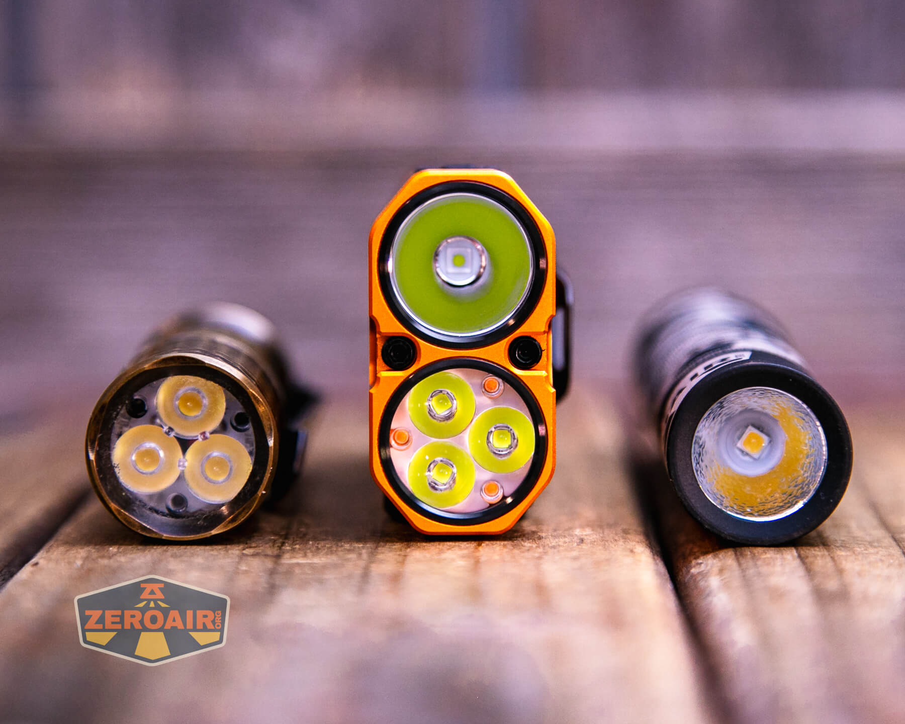Three flashlights are standing upright on a wooden surface, viewed from the front. The flashlight in the center is orange with four LED emitters, and the other two have three and one emitters, respectively. A ZeroAir logo is visible.