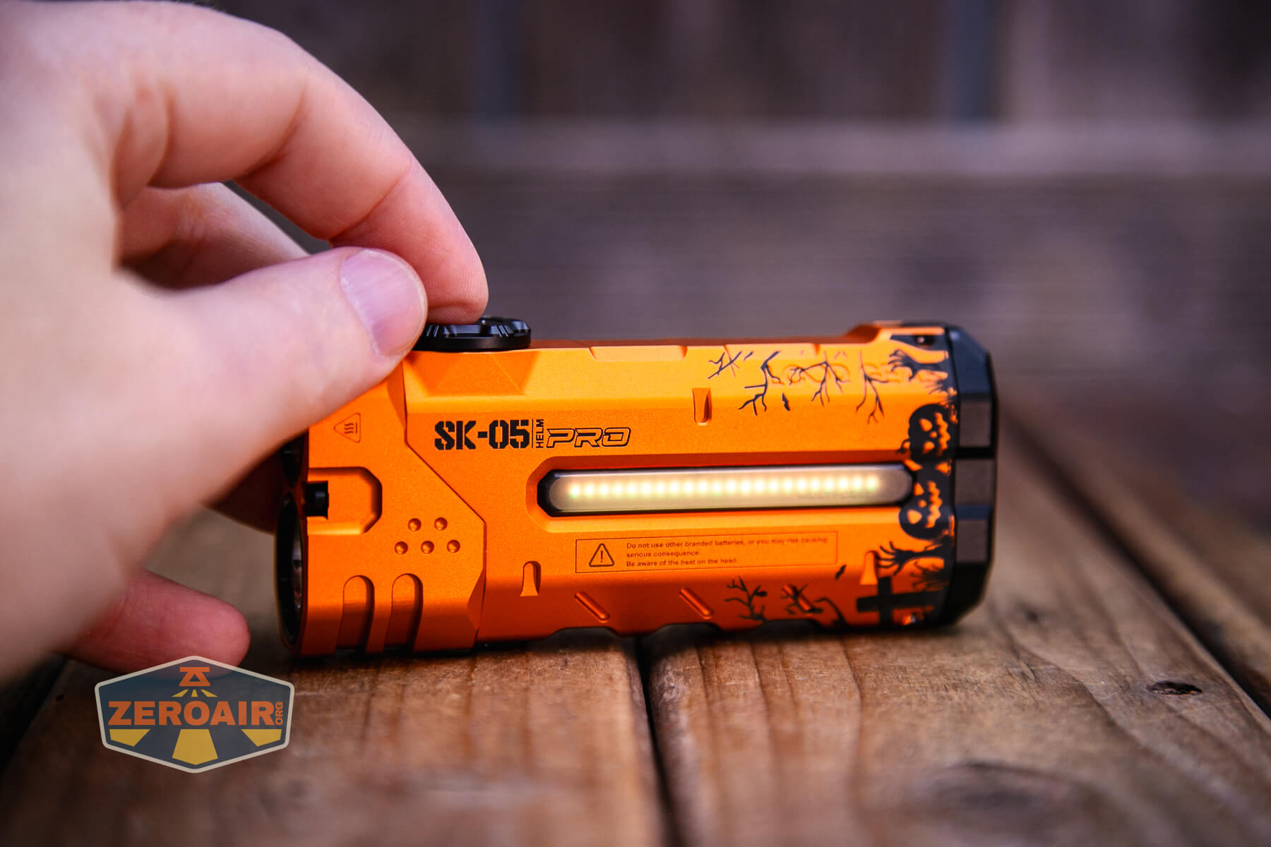 A hand holds an orange flashlight labeled SK-051EPRO with a horizontal LED strip, black designs, and a side switch, placed on a wooden surface. A ZEROAIR logo sticker is also visible on the surface.