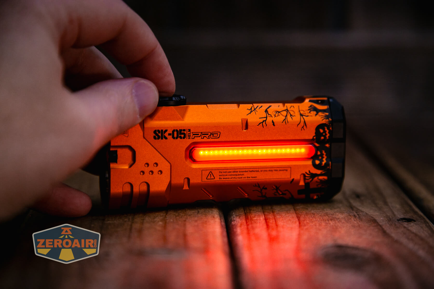 A hand holds an orange SK-051 PRO device with a lit red LED strip, resting on a wooden surface. The device has various markings and a logo reading ZeroAir in the corner.