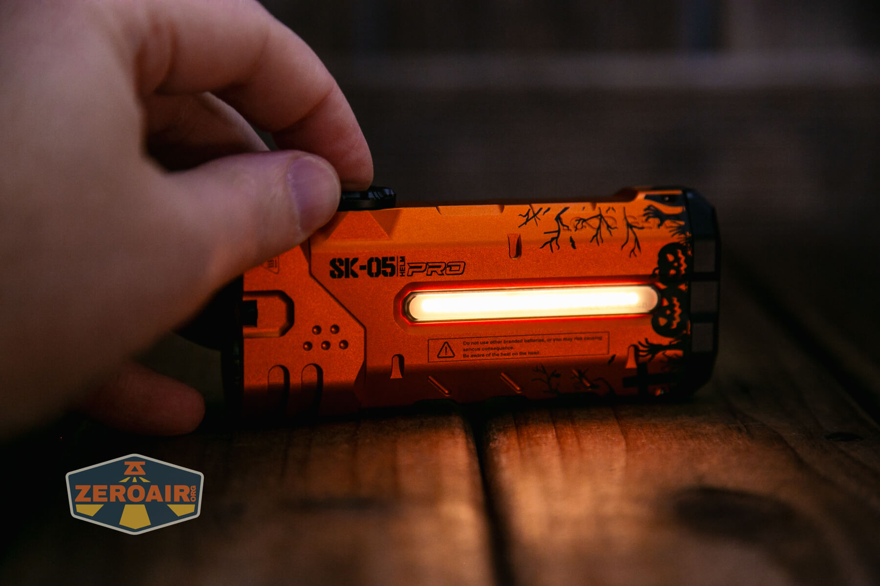 A left hand holds an orange SK-051 PRO flashlight with a lit side panel on a wooden surface. The flashlight has black decorative accents, and a ZeroAir logo is visible in the bottom left corner.
