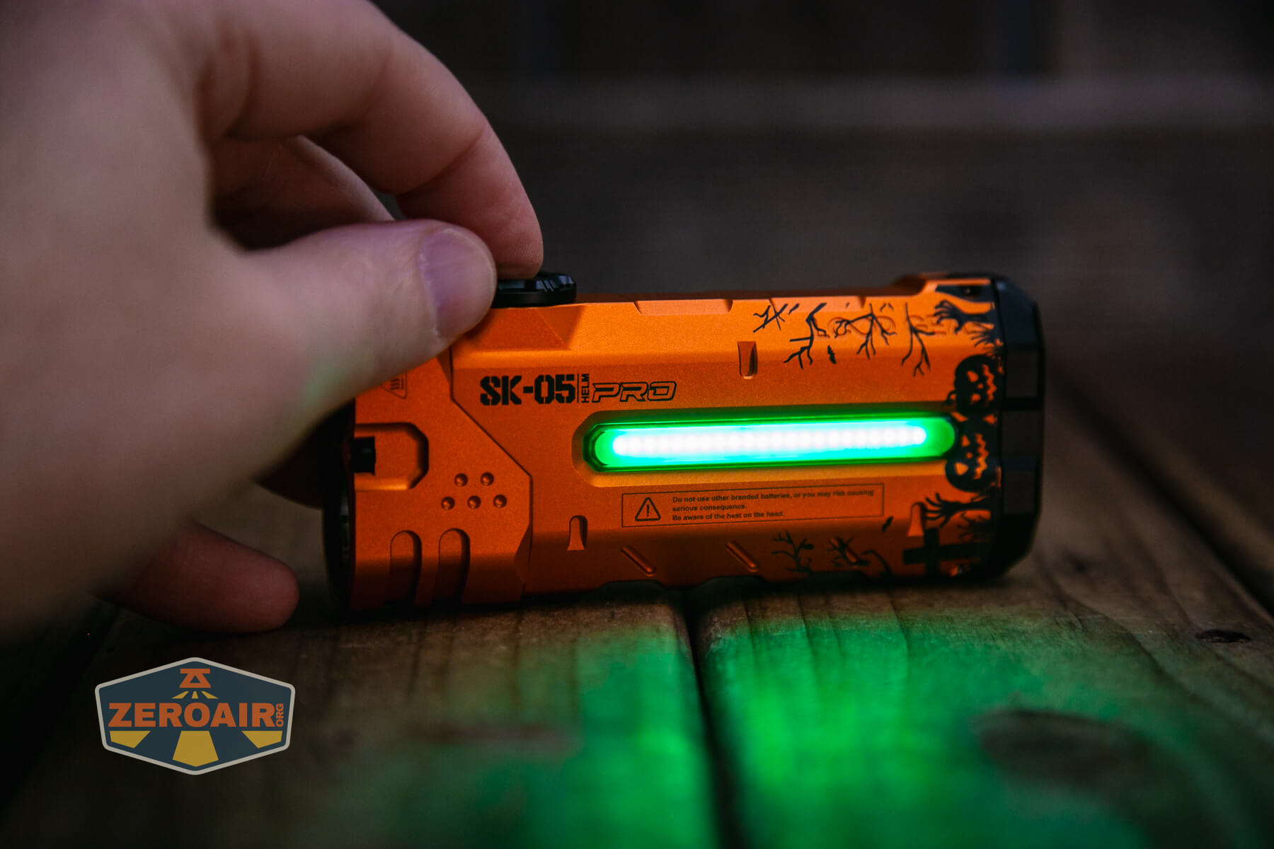 A hand holds an orange SK-05i PRO flashlight with a glowing green light strip, on a wooden surface. The flashlight features black decorative patterns and a warning label. A ZeroAir logo is visible in the bottom left corner.