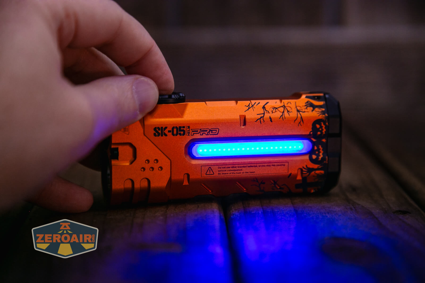 A hand holds an orange SK-051 PRO device with a horizontal blue LED light strip illuminated on its side, resting on a wooden surface. A ZeroAir logo is visible in the lower left corner.