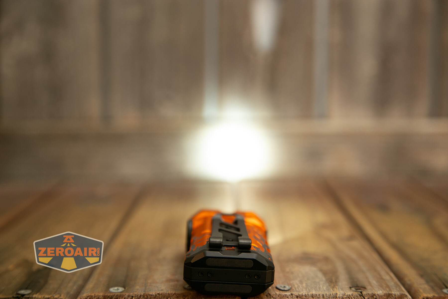 A small orange flashlight is turned on and lying on a wooden surface, shining a bright beam of light toward a wooden wall. A ZeroAir logo is visible in the bottom left corner.