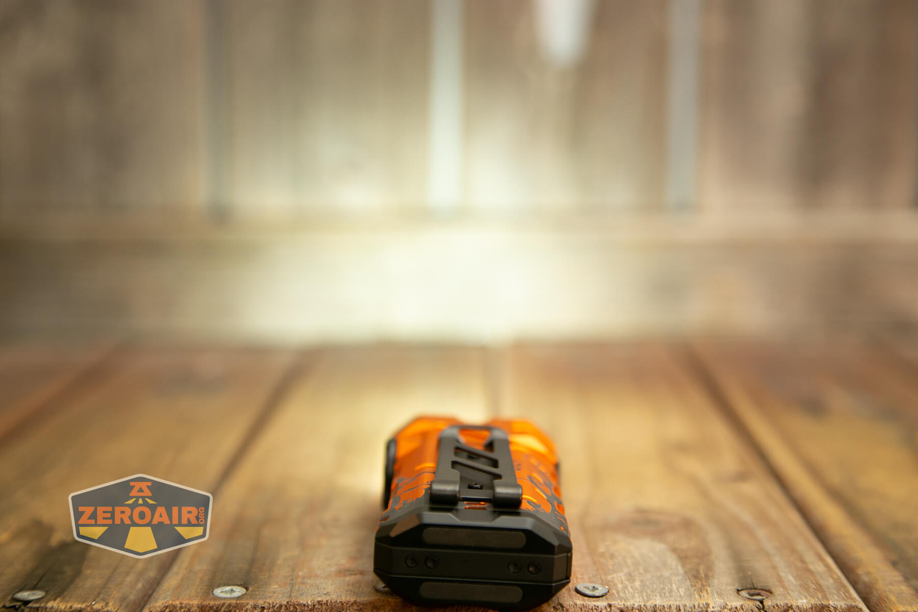 An orange and black flashlight is lying on a wooden surface, facing away from the camera. The background is out of focus, and a ZEROAIR! logo appears in the bottom left corner.