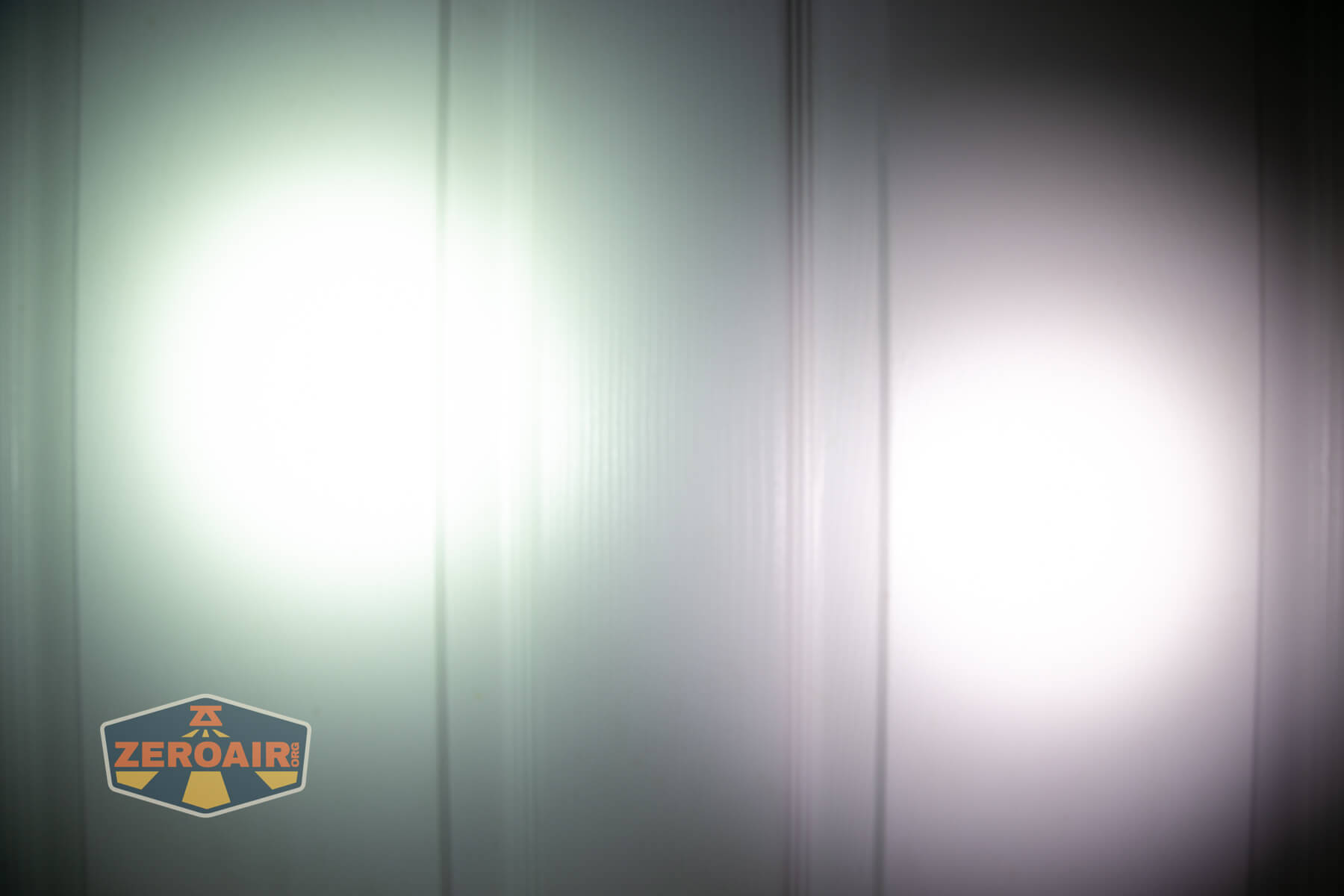 Two bright flashlight beams, one with a greenish tint on the left and one with a whiter tint on the right, shine onto a flat vertical surface. There is a ZerOair logo in the bottom left corner.